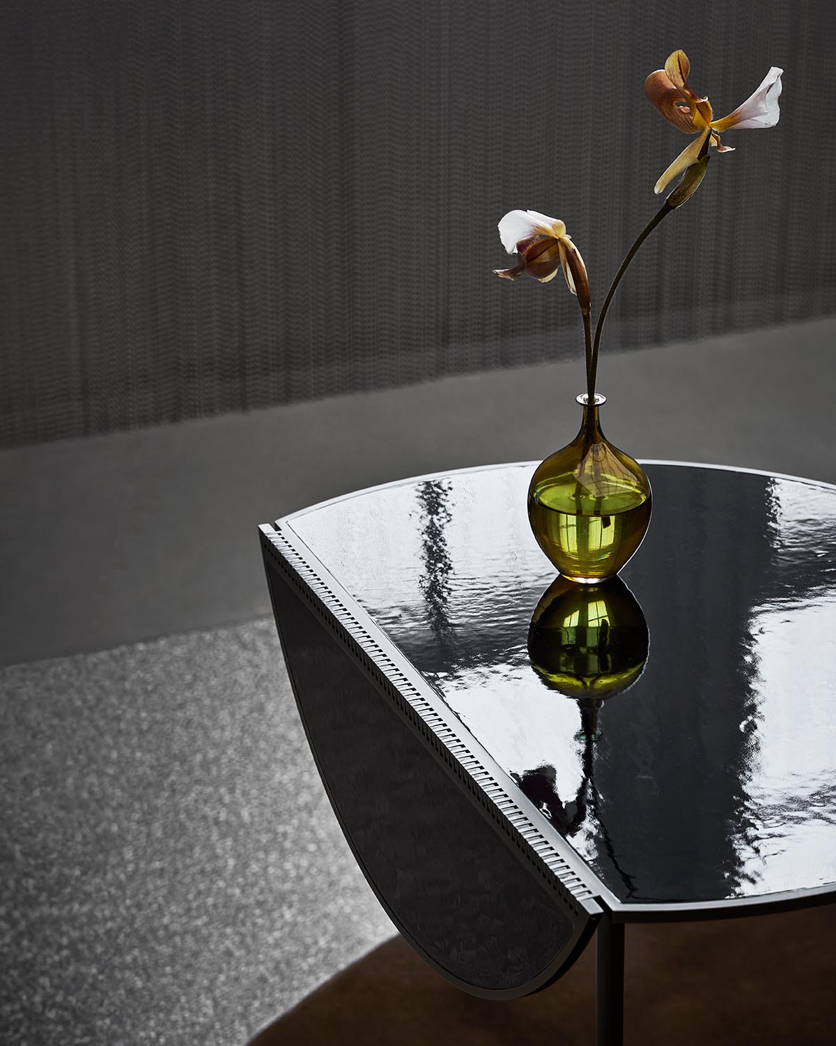 Close-up of a glossy black bistro table with rounded edges and a green vase holding two delicate flowers on top