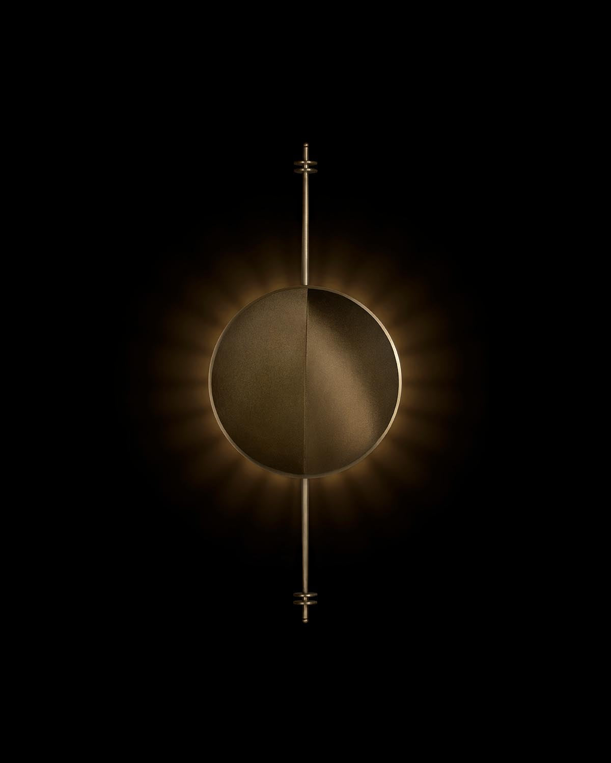 Round bronze wall light with vertical rod and illuminated halo against black background