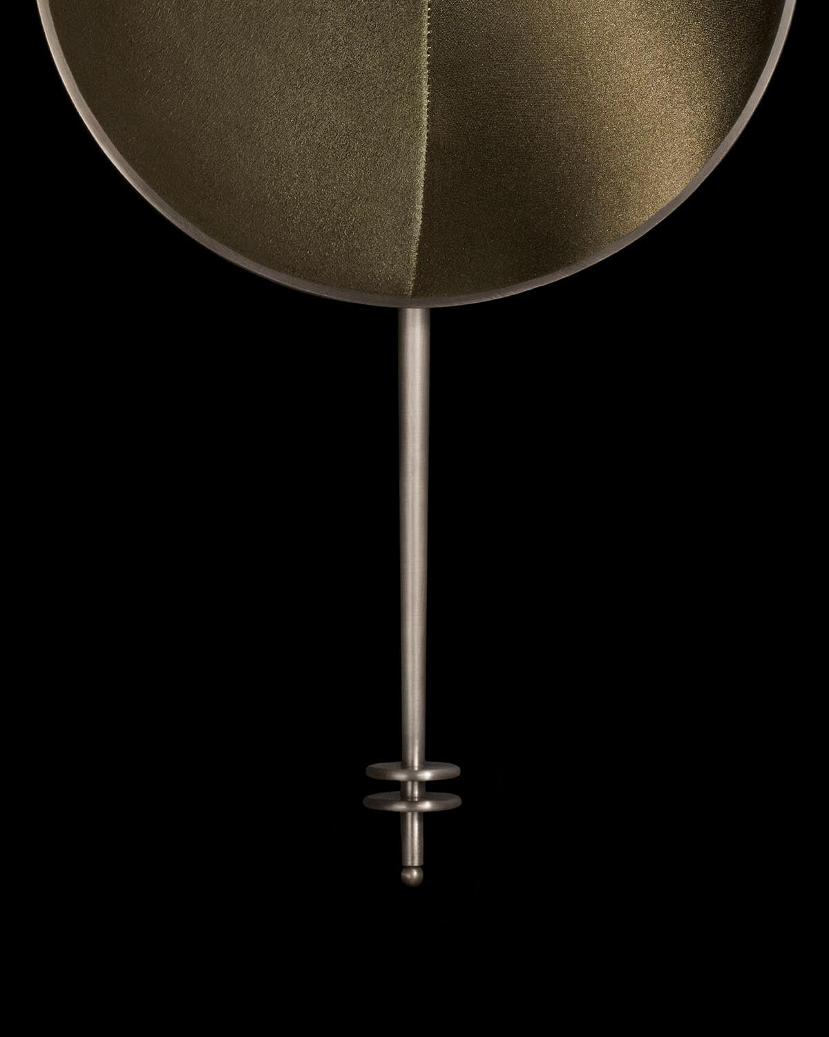 Close-up of a minimalist metal wall sconce with a circular textured backplate and slender vertical rod in warm tones against black background