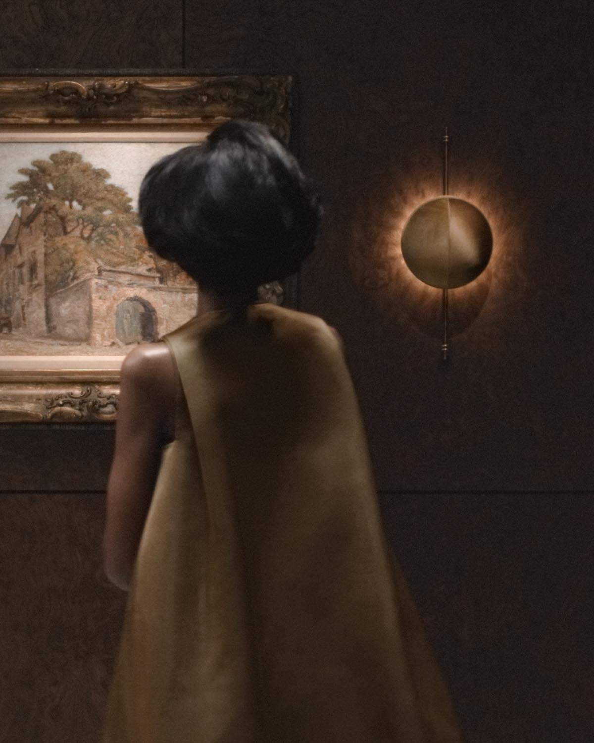 Dimly lit wall sconce with circular metallic design casting a warm glow beside a framed painting in a gallery setting