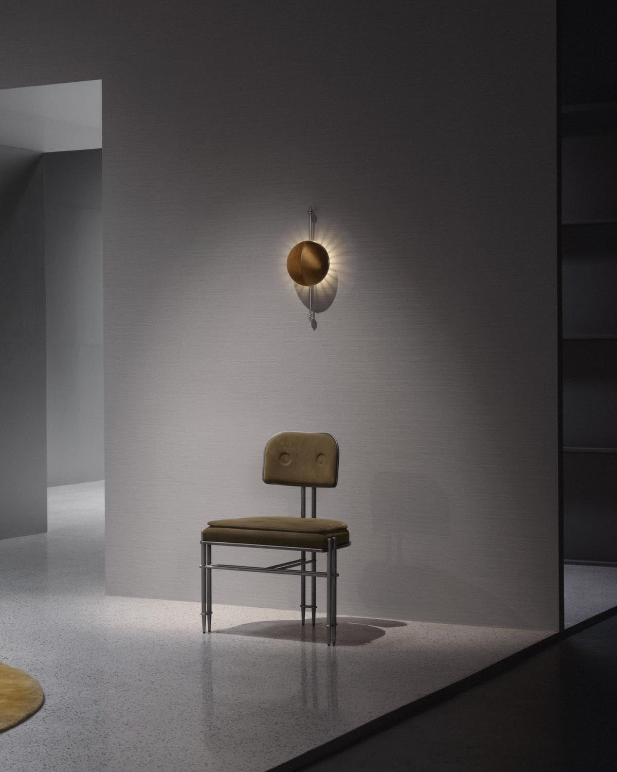 Wall-mounted round bronze sconce casting radial light above a minimalist chair with olive green cushions against a gray wall