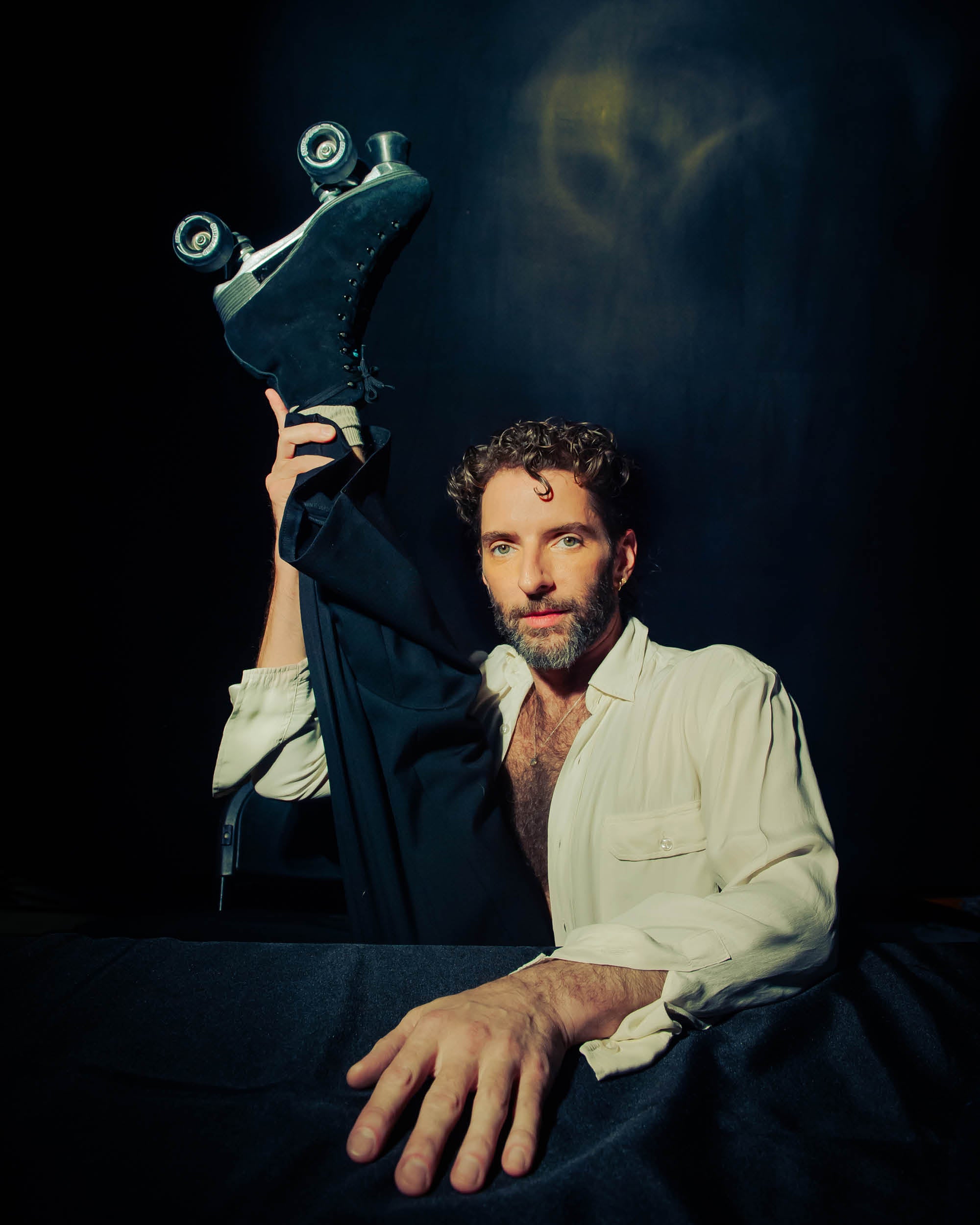 Man with curly hair and beard posing with one leg raised, wearing black roller skates and a white shirt against dark background