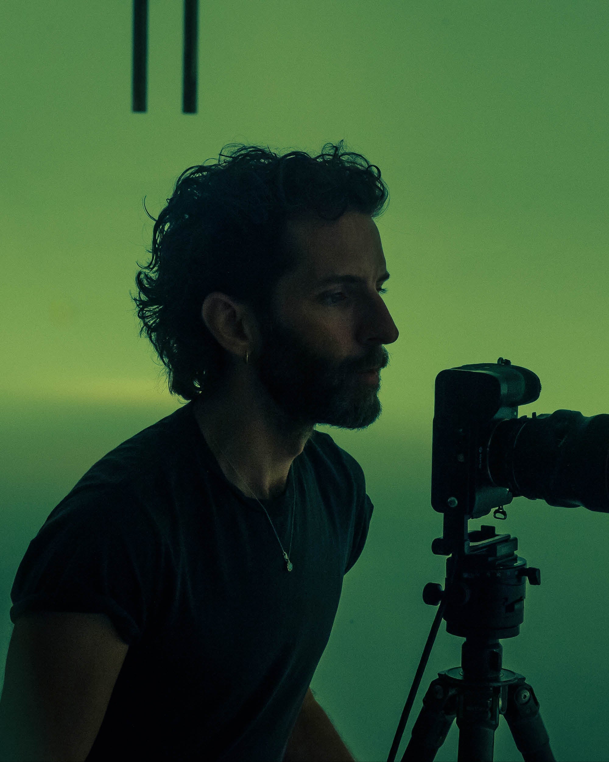 Side profile of a man with curly hair and beard focused on a camera mounted on a tripod against a green background