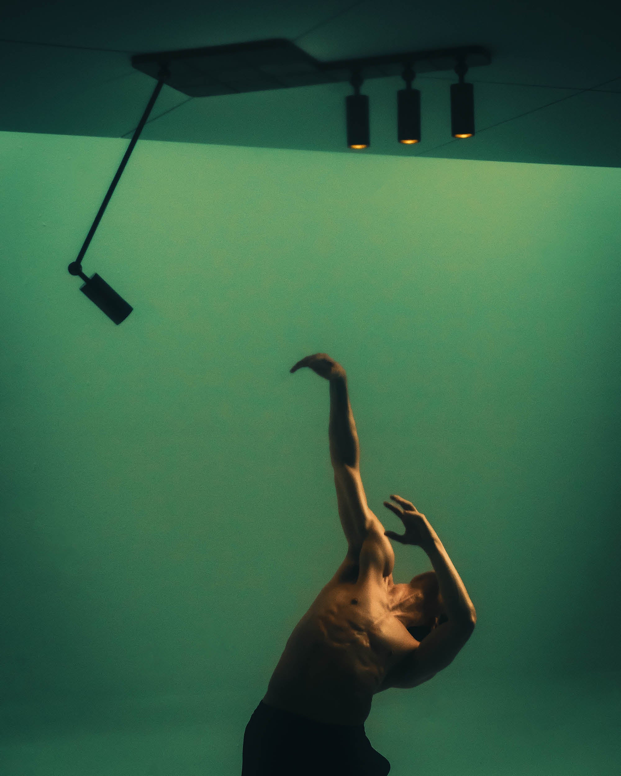 Single shirtless dancer with muscular build posed with arms raised against a green gradient background under ceiling lights