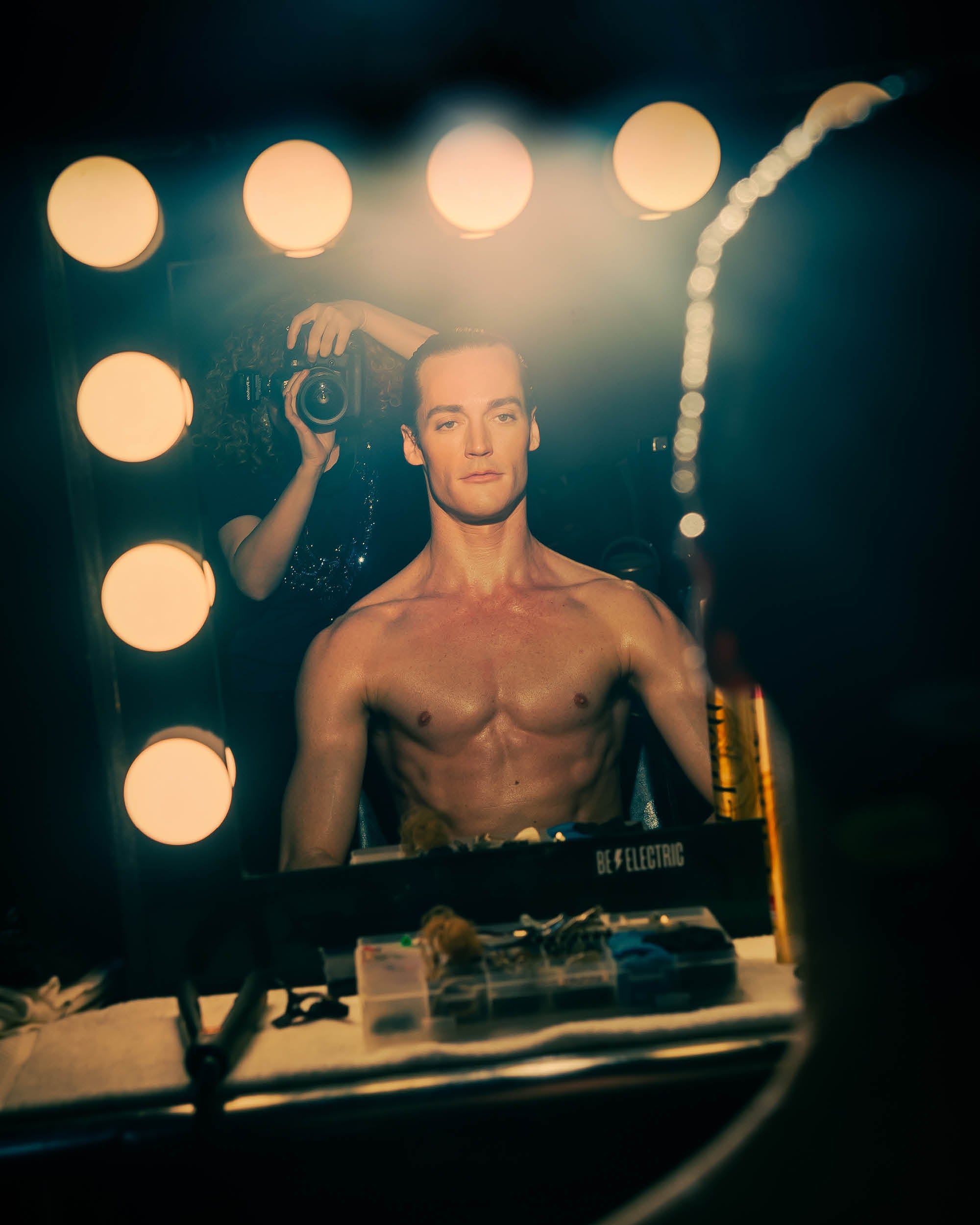 Shirtless man looking into a vanity mirror framed by illuminated round bulbs with a photographer reflected behind him