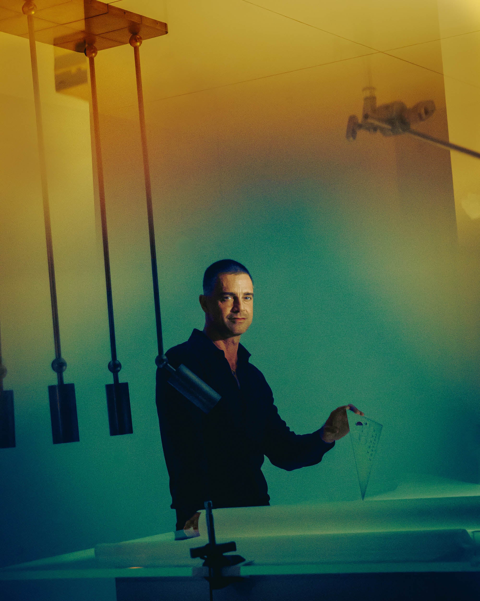 Man in dark clothing holding a translucent triangular ruler in a dimly lit room with hanging weights and scientific equipment