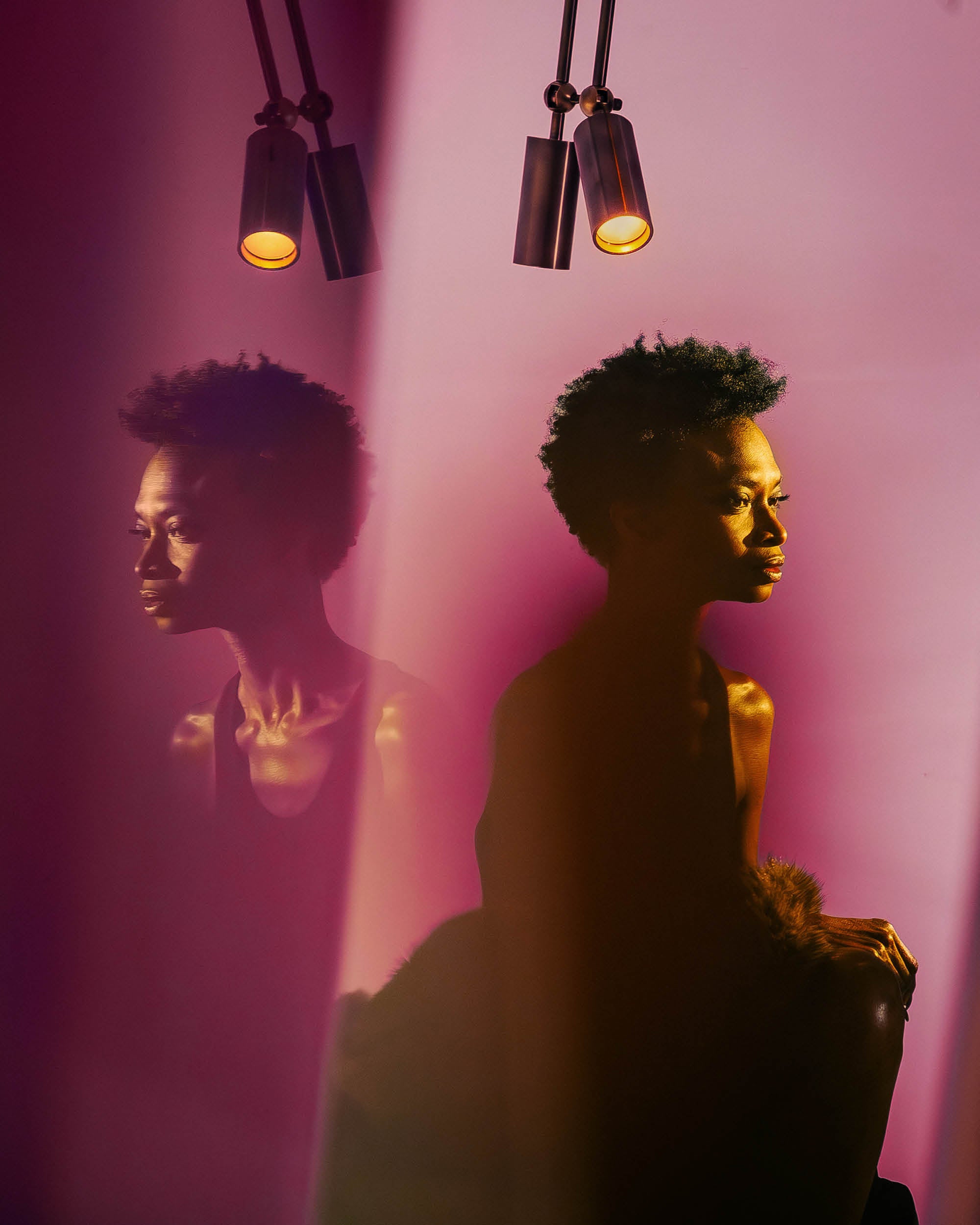 Side profile of a woman illuminated by warm light against a pink background with her reflection visible nearby