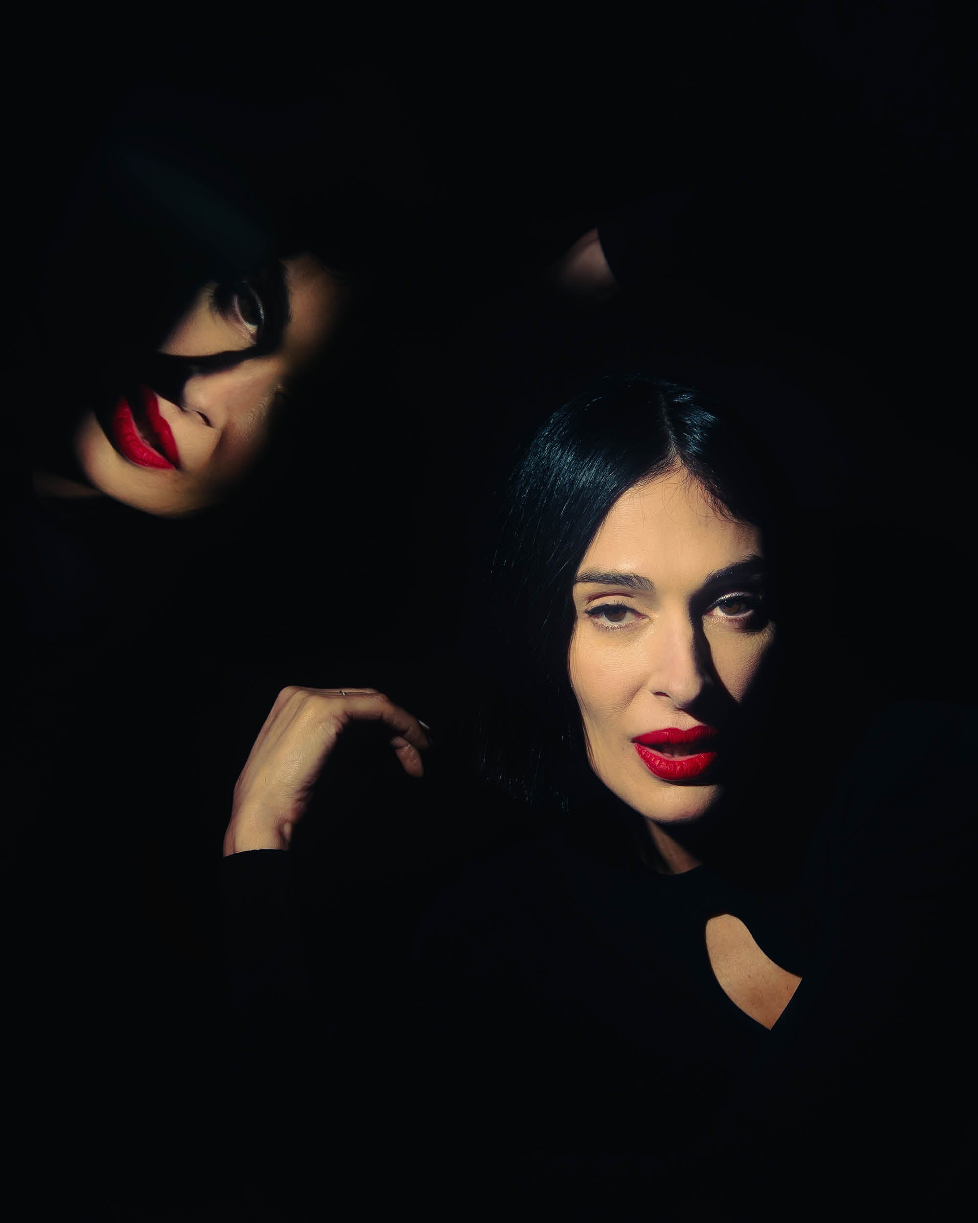 Two women with black hair and red lipstick partially illuminated against a dark background with dramatic shadows