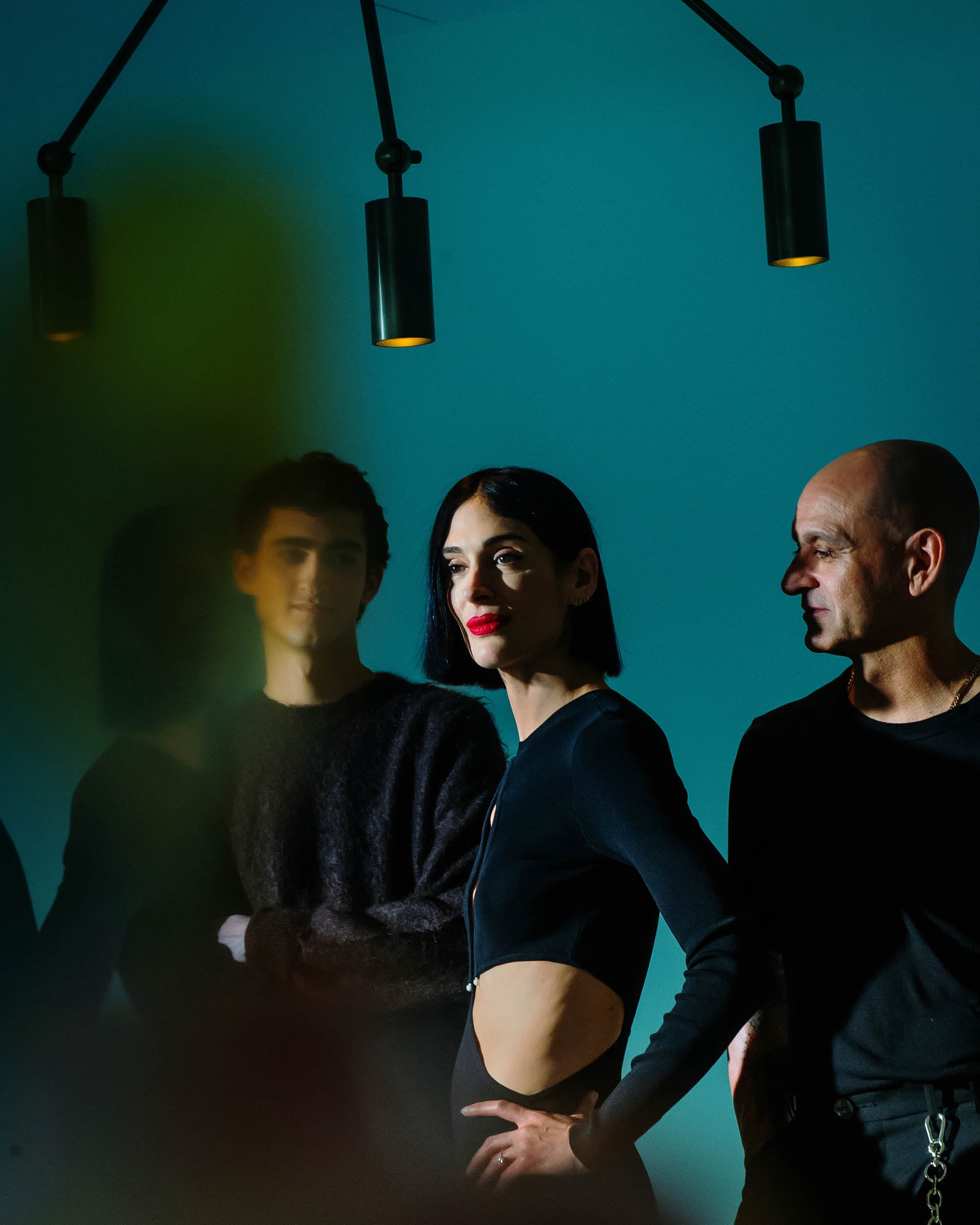 Group of three people with focused expressions standing under three black pendant lights against a teal background
