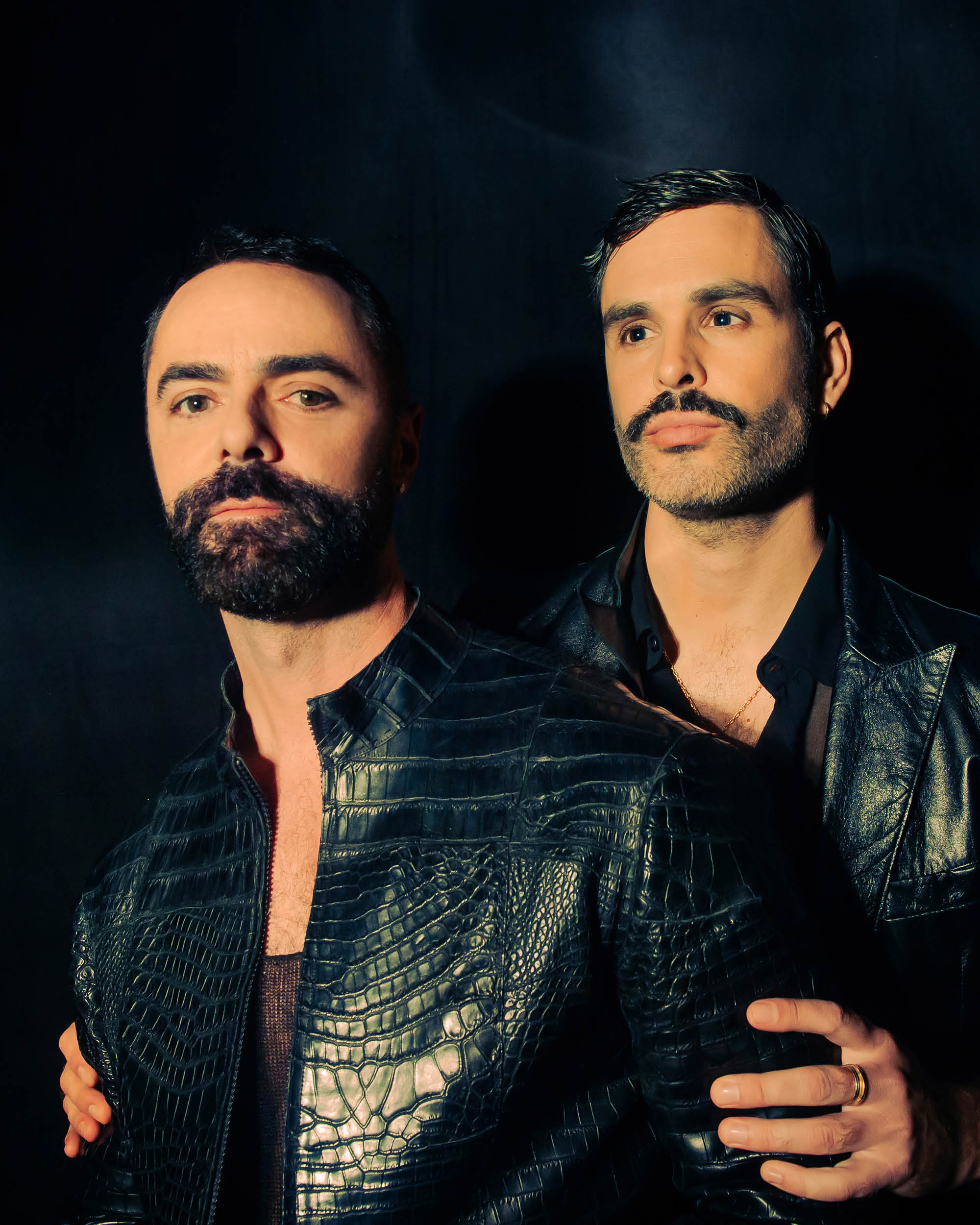 Portrait of two men with beards wearing dark textured leather jackets against a black background