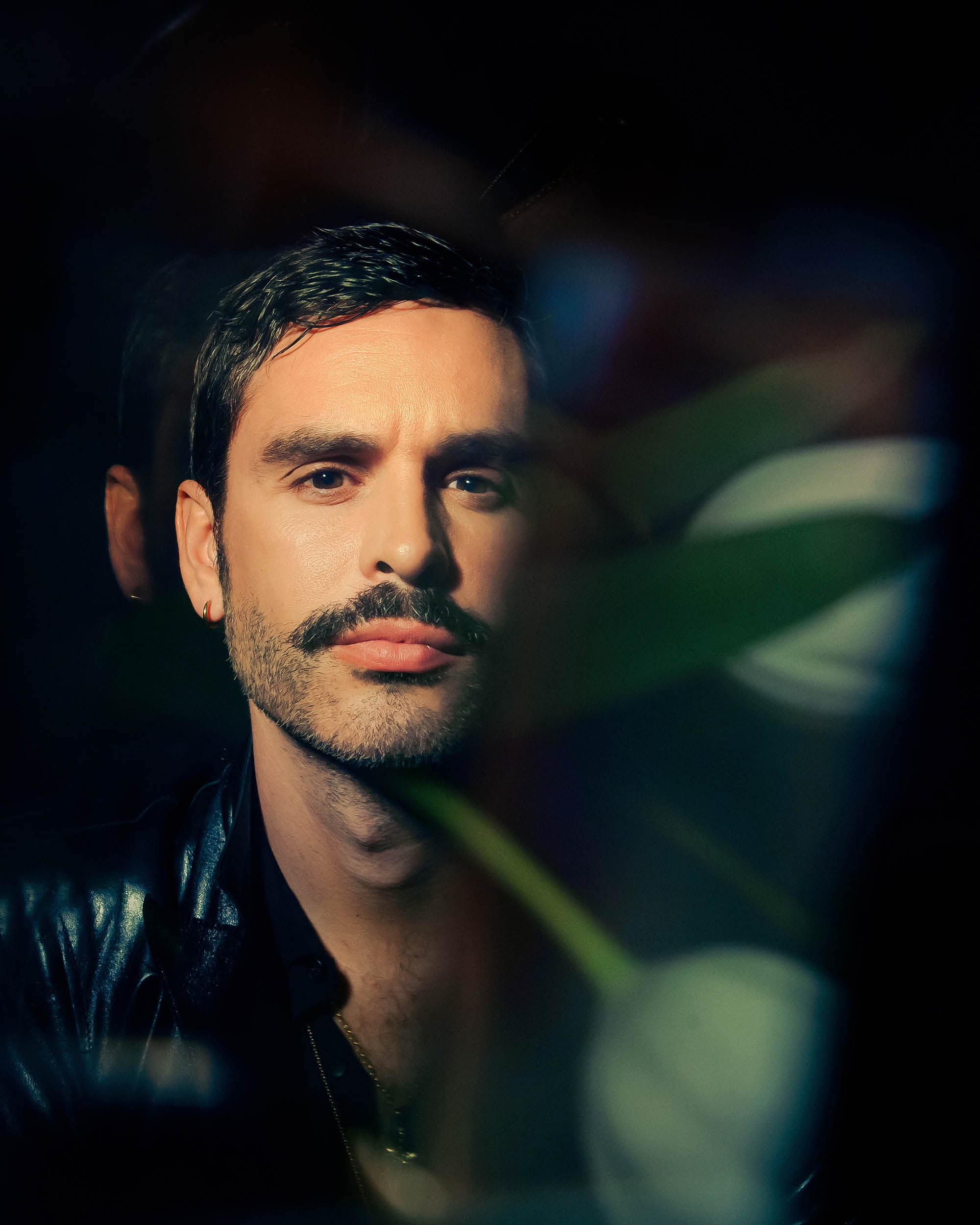 Close-up portrait of a man with a mustache and short hair, partially obscured by soft focus foliage in dark lighting