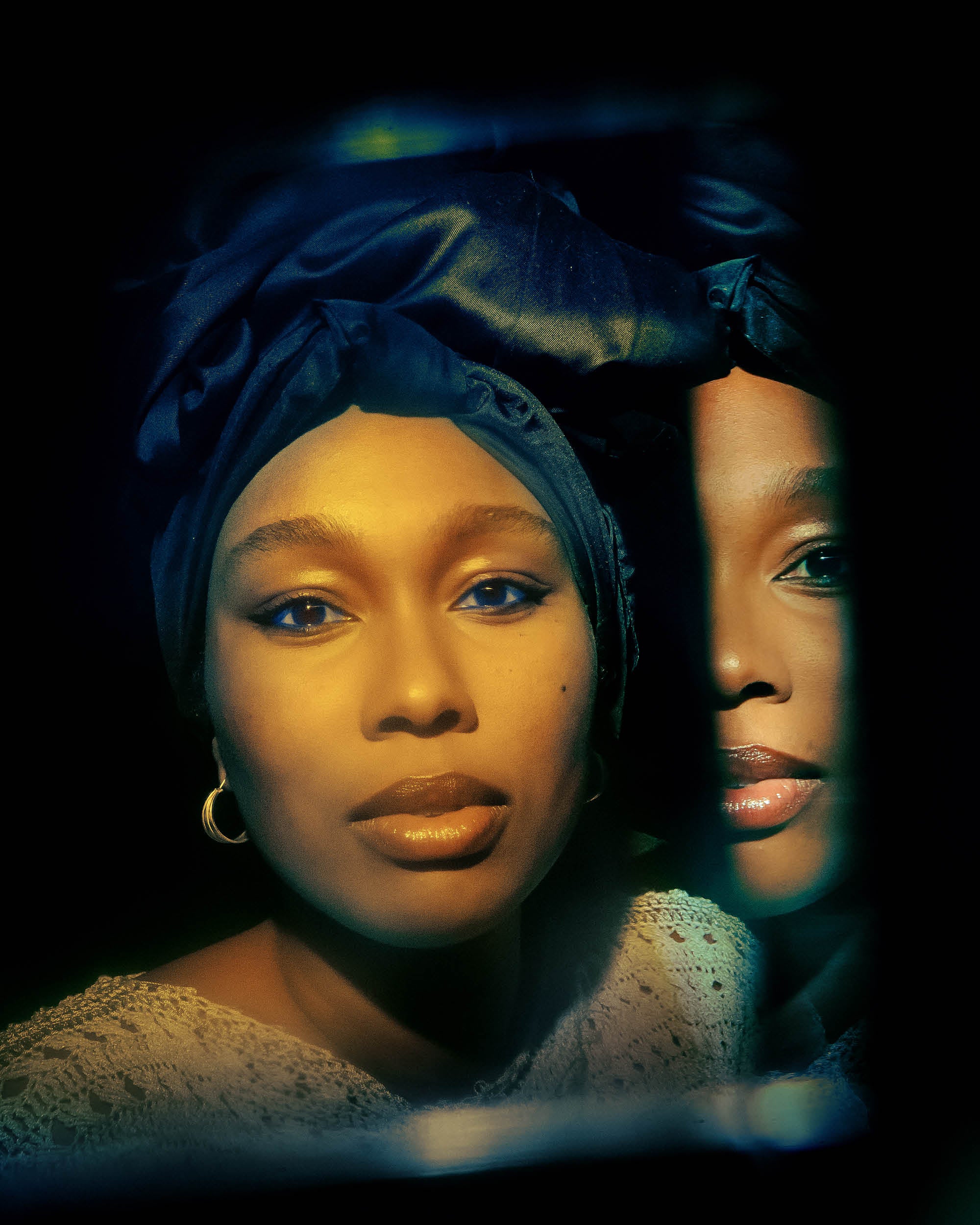 Portrait of a woman wearing a dark headwrap and gold hoop earrings reflected in a mirror against a dark background