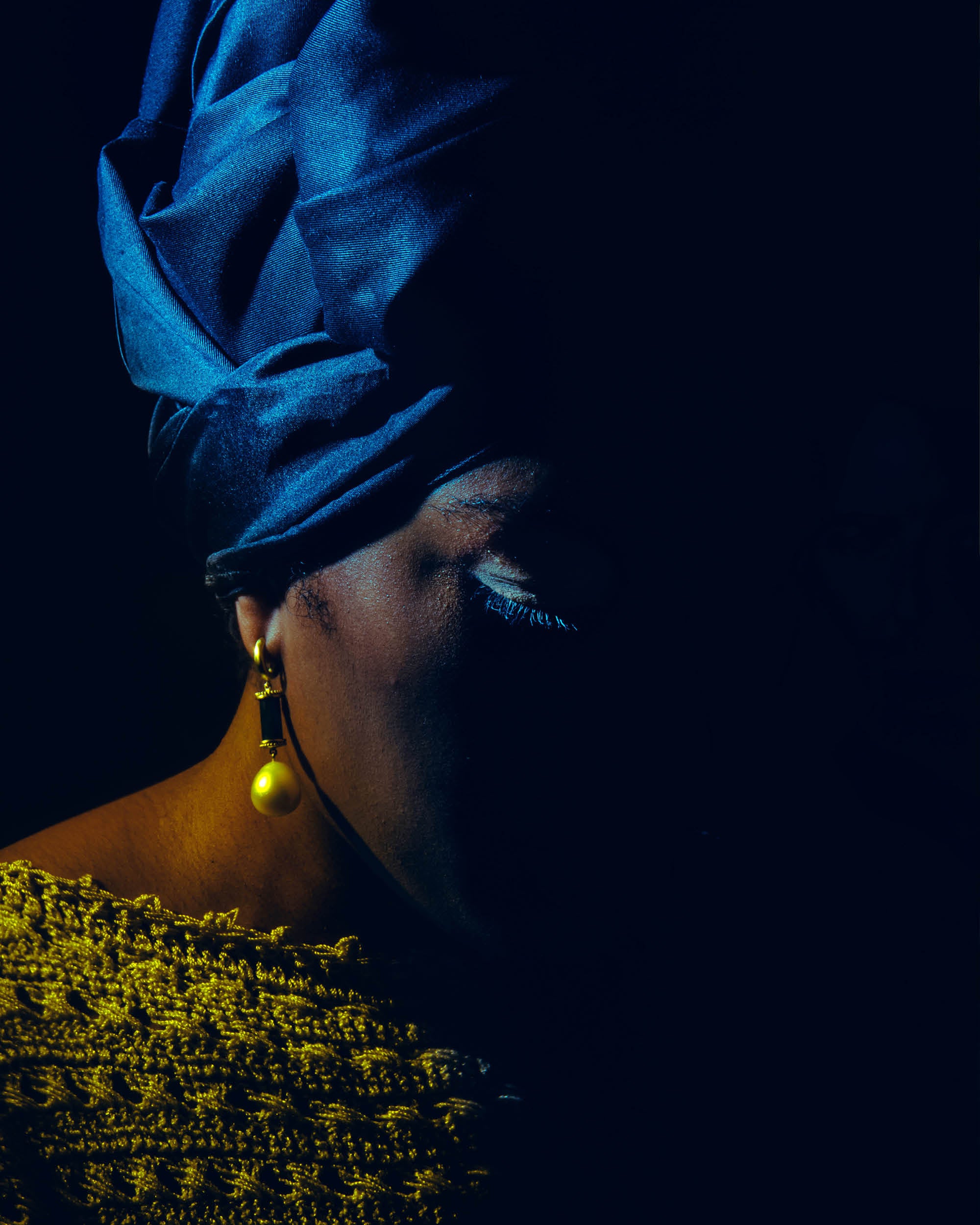 Portrait of a woman in shadow wearing a blue headwrap, gold pearl earring, and textured yellow garment against black background