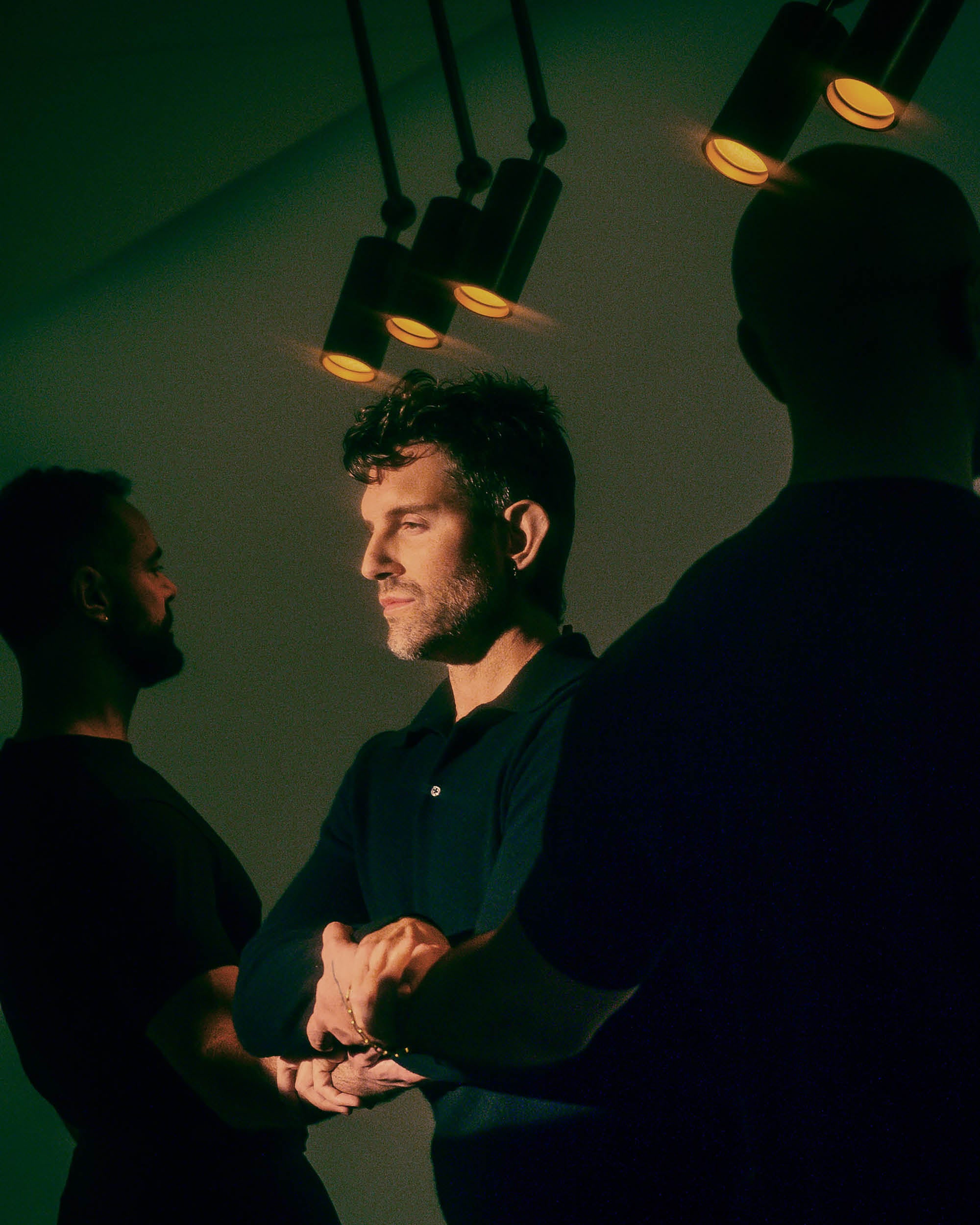 Three men in dark clothing standing close together under warm overhead lights in a dimly lit room