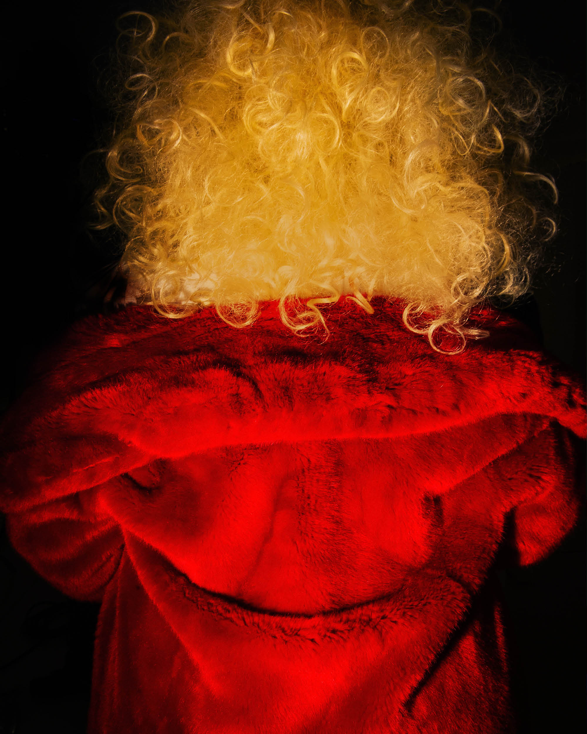 Back view of a person with voluminous curly blonde hair wearing a bright red plush jacket against a black background