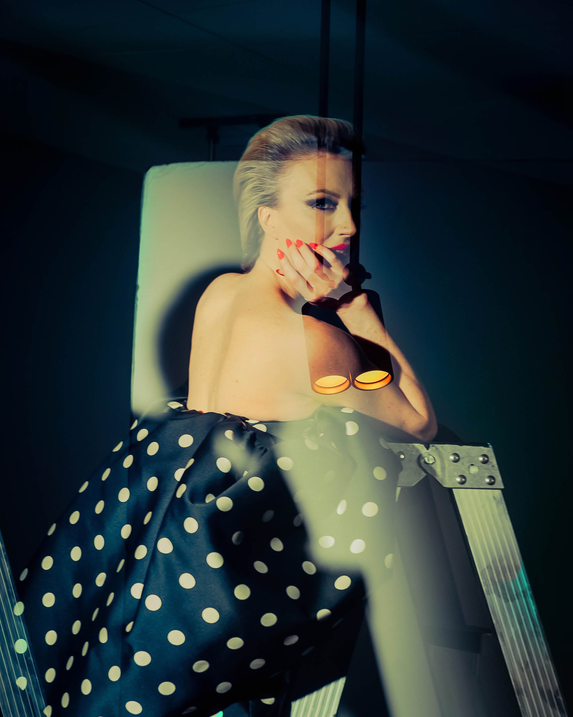 Woman with styled hair and red nails wearing a black and white polka dot skirt leaning on a metal ladder in dim lighting