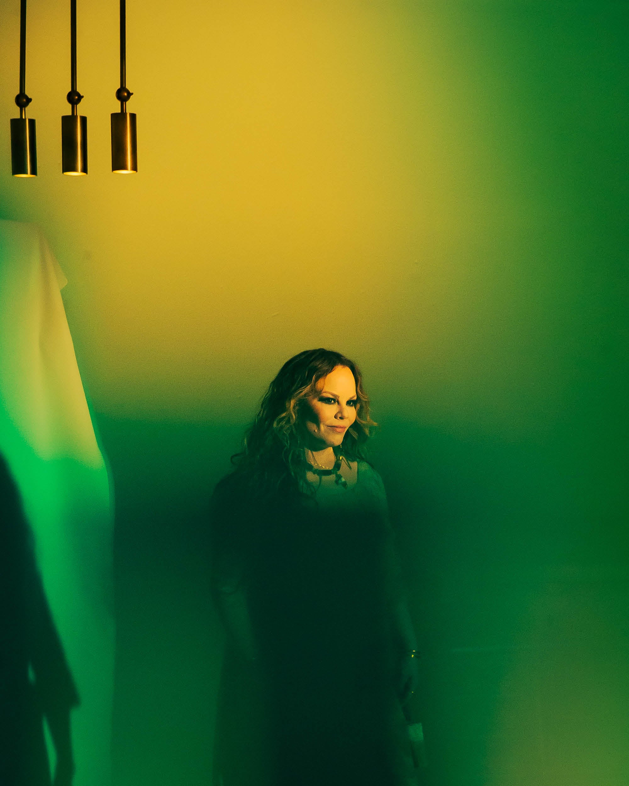 Woman with wavy hair standing in green and yellow gradient light under three cylindrical pendant lights
