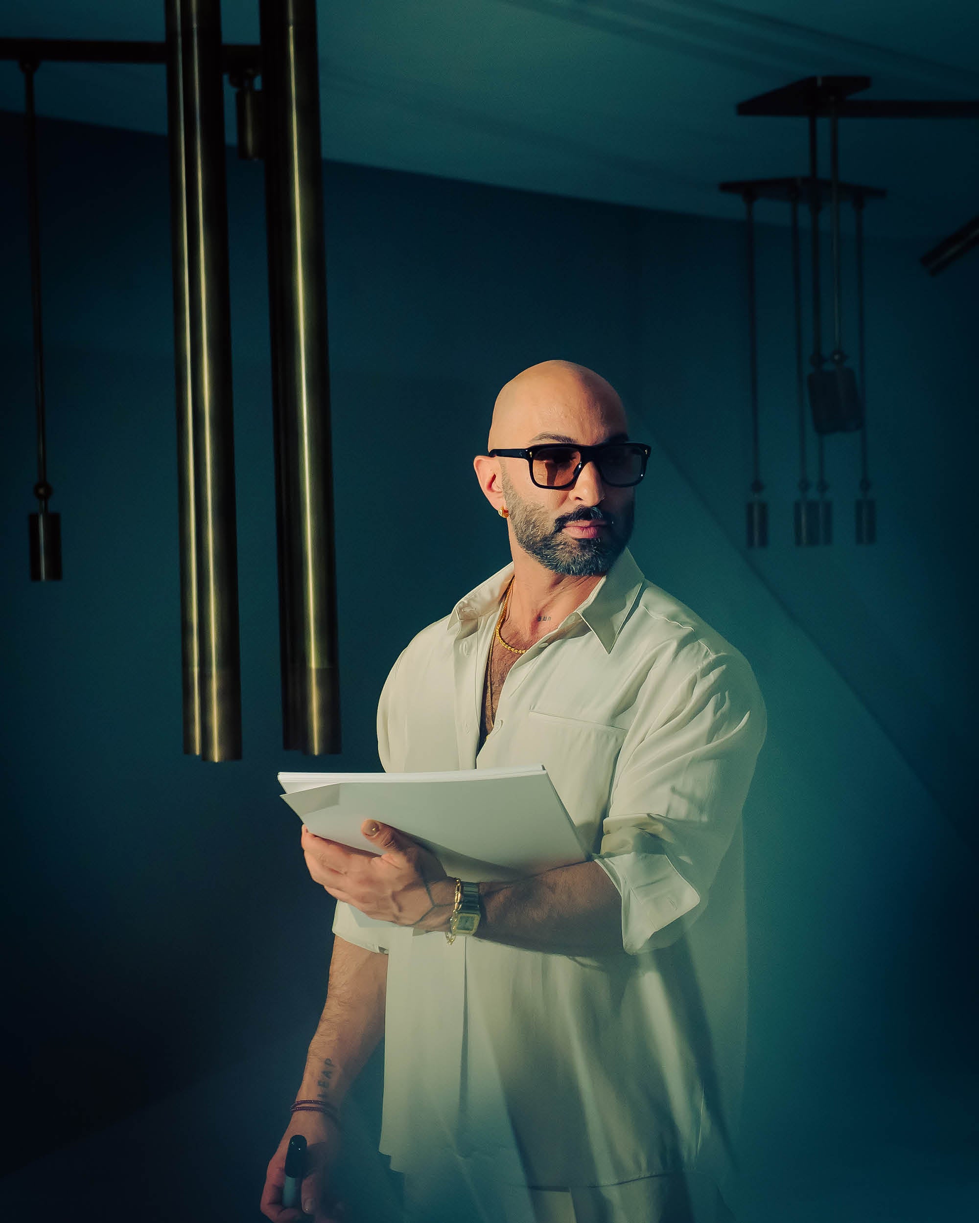 Man wearing dark sunglasses and a white shirt holding papers in a dimly lit modern interior space