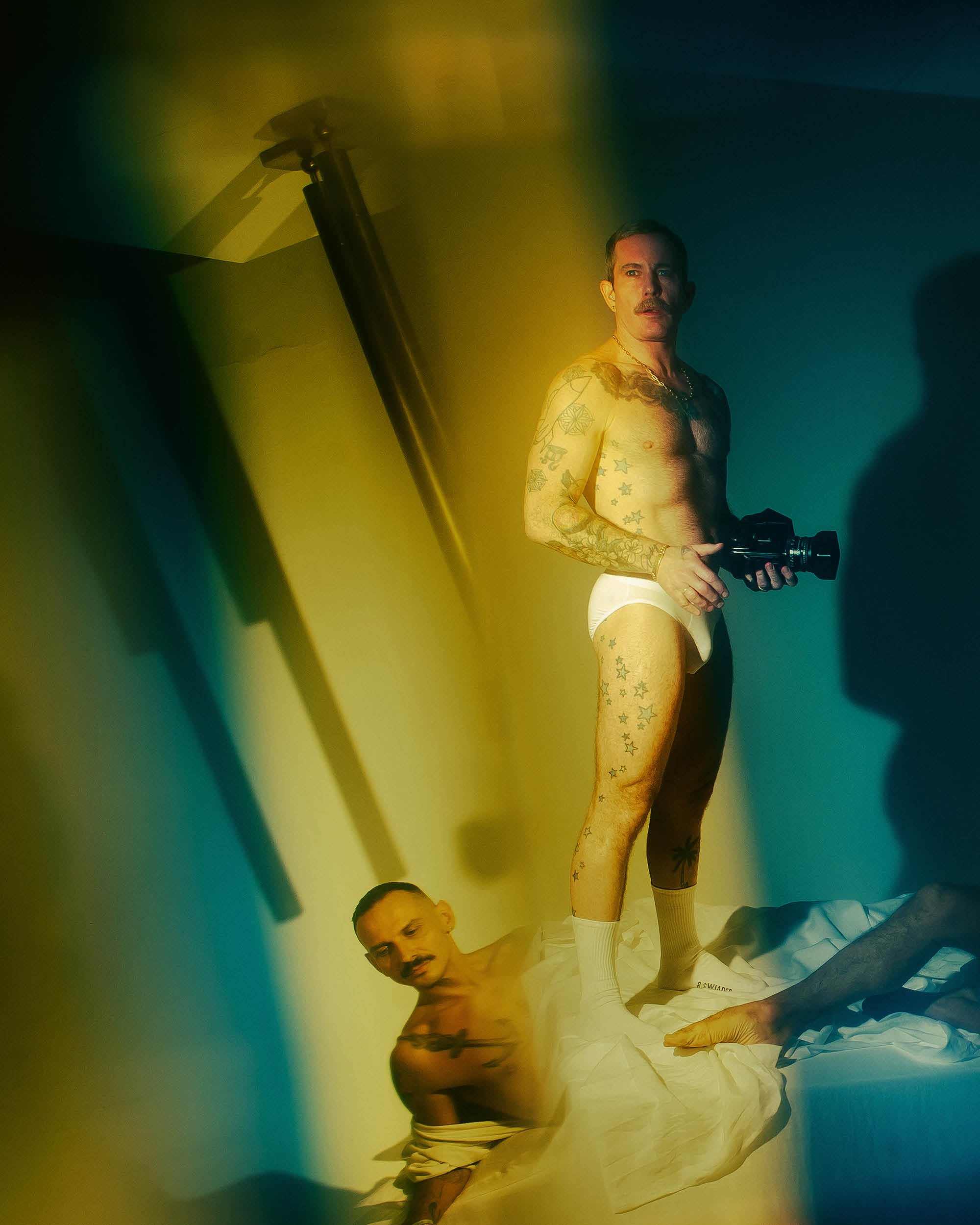 Artistic photograph of two tattooed men in white underwear and socks, with dramatic shadows and blurred yellow lighting effects