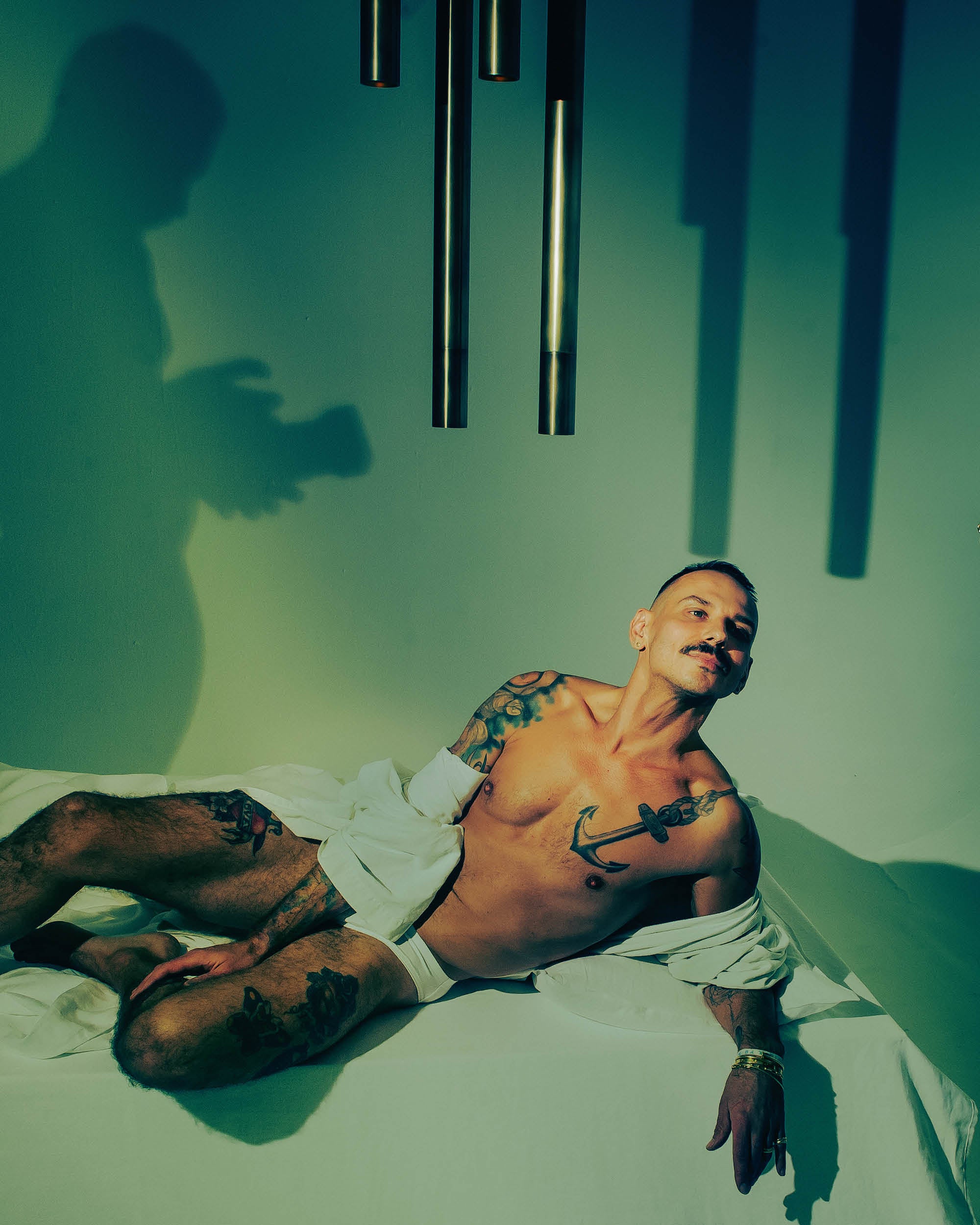 Shirtless man with tattoos reclining on white surface under greenish light with shadows and hanging metal tubes above