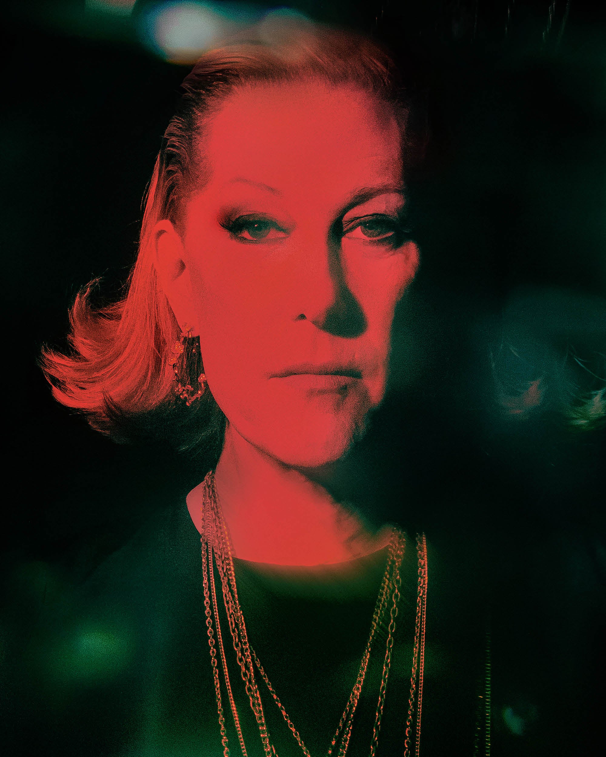 Portrait of a person with short hair wearing layered gold chains and earrings, illuminated by red and green lighting on a dark background