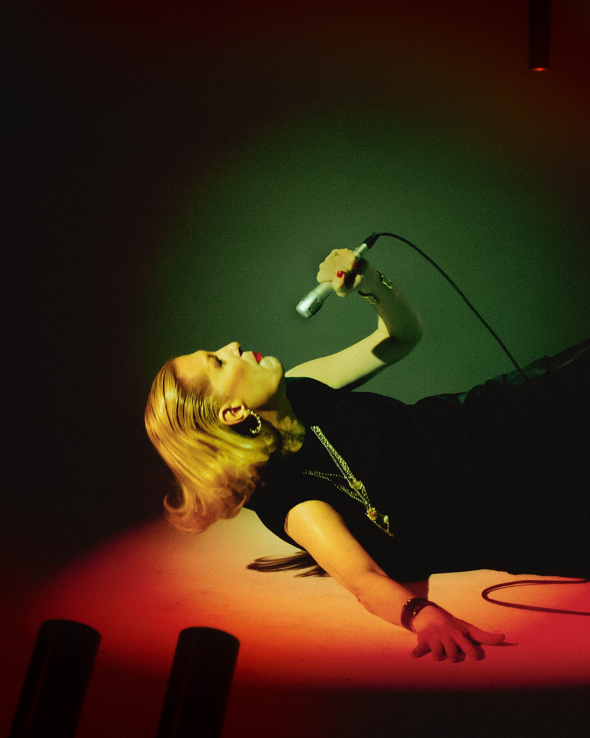 Side view of a woman with slicked-back hair lying on the floor singing into a microphone under colorful lighting