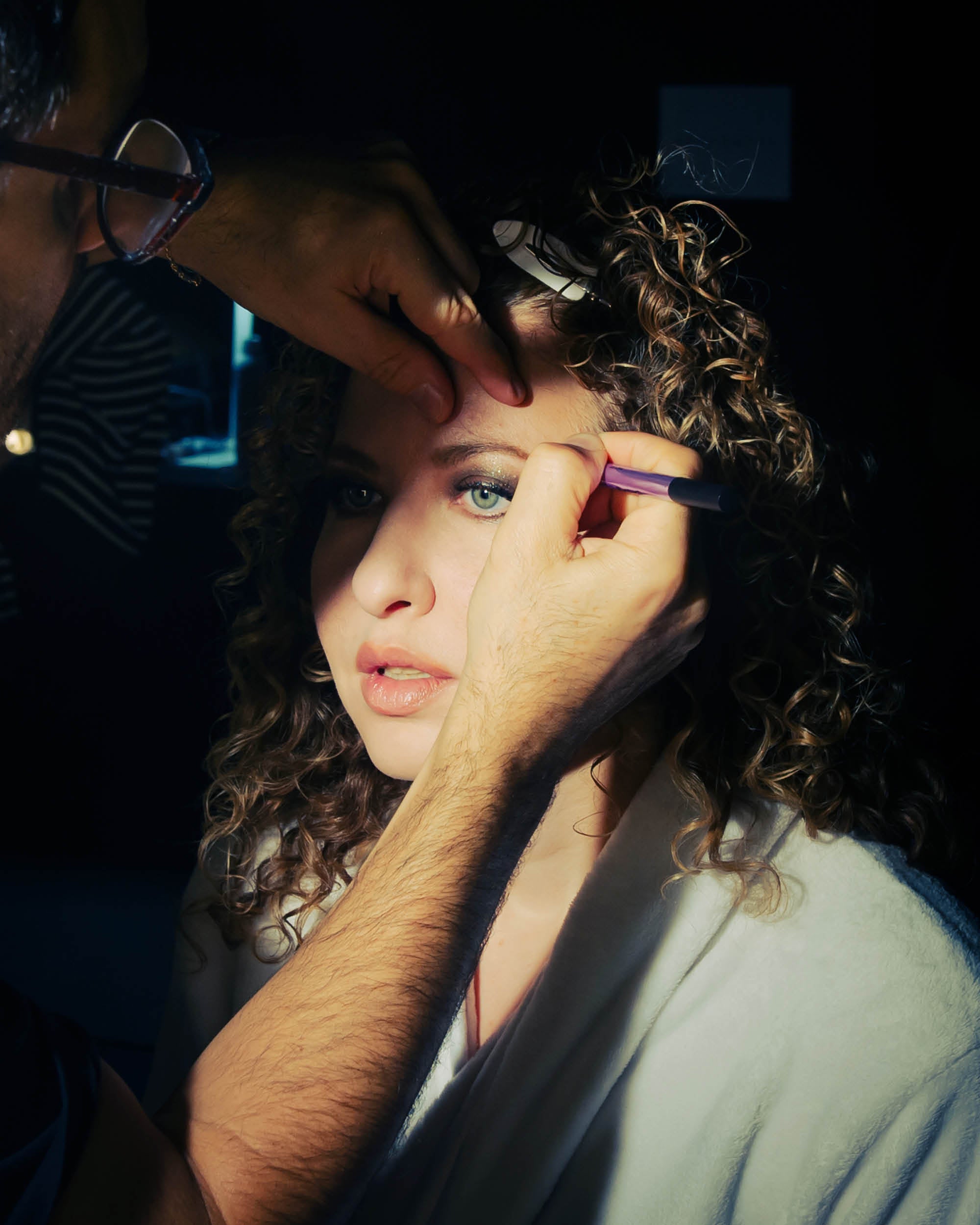 Close-up of a woman with curly hair having makeup applied to her eyebrow by a person in a dimly lit setting