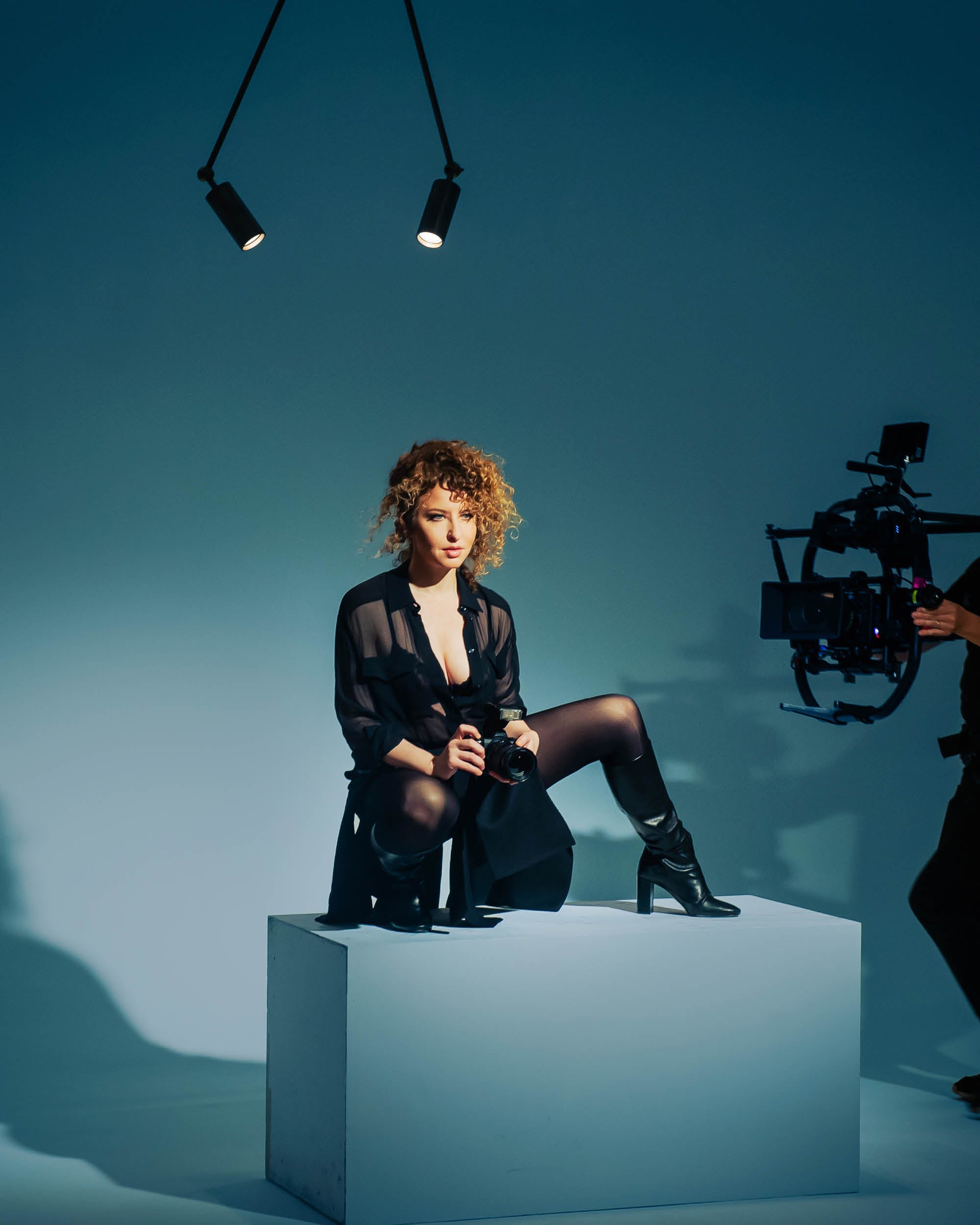 Woman with curly hair in sheer black outfit sitting on white cube holding a camera under overhead studio lights