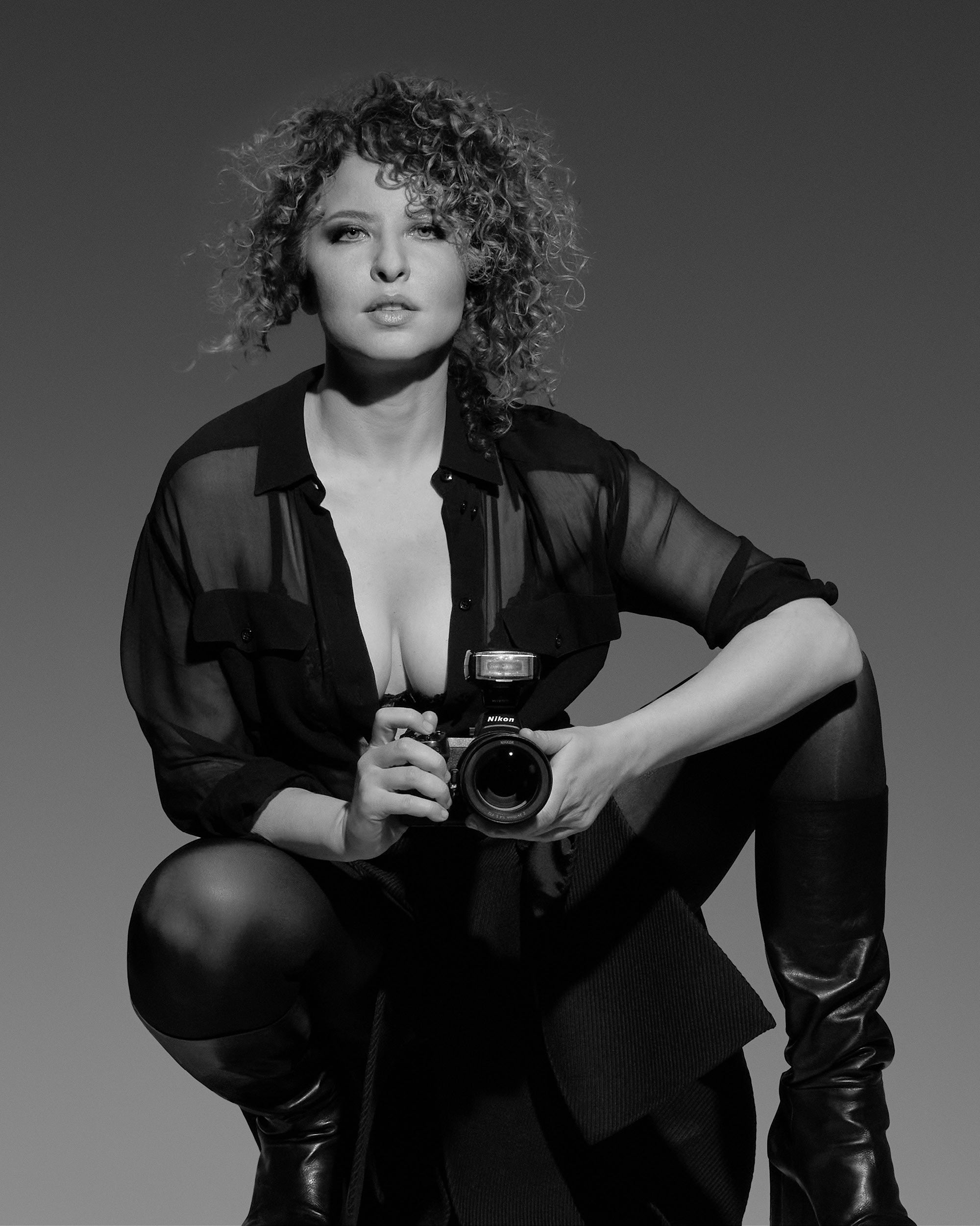 Black and white portrait of a curly-haired woman wearing a sheer shirt and boots, holding a camera and looking forward