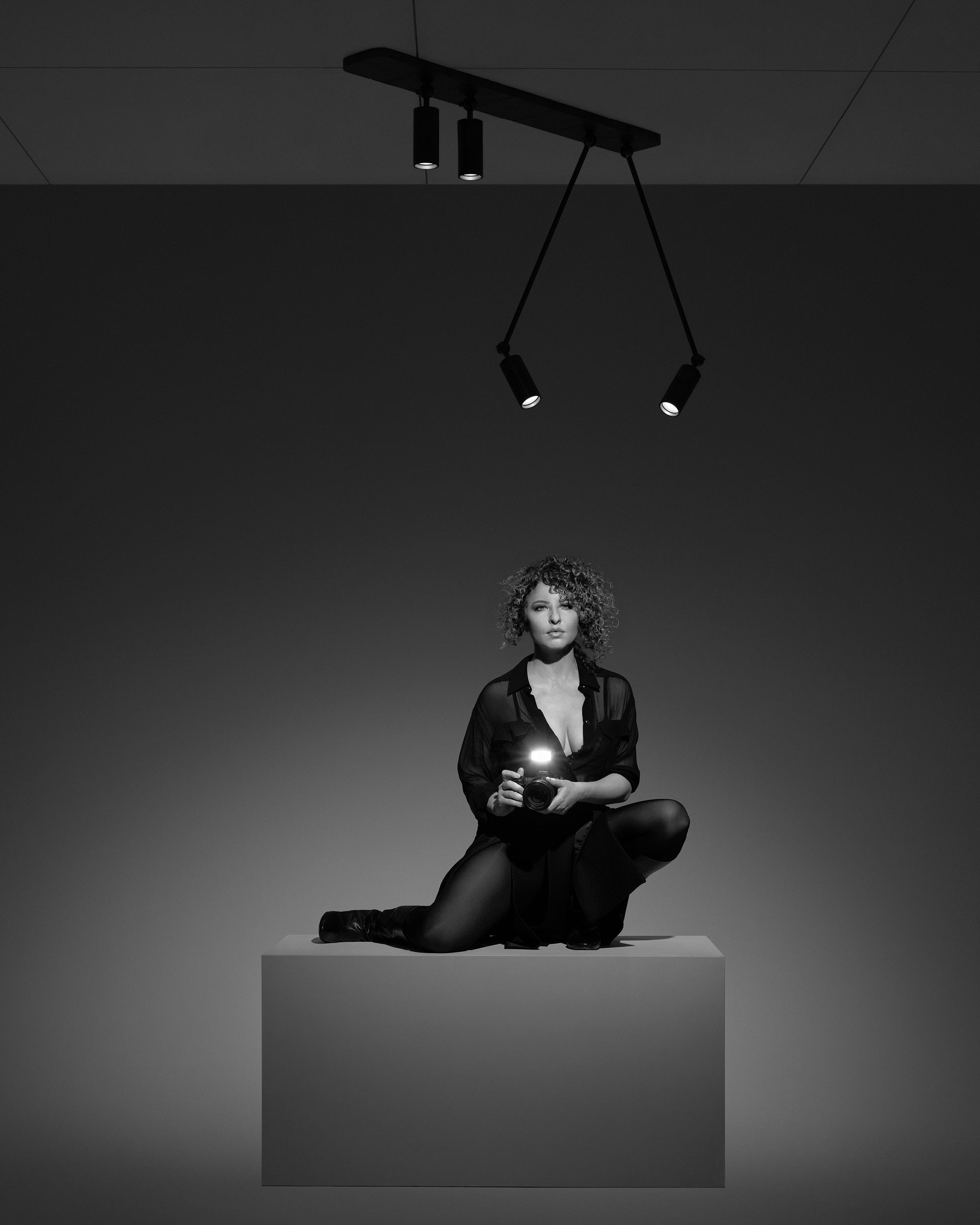 Black and white photo of a woman with curly hair kneeling on a pedestal holding a camera under spotlights