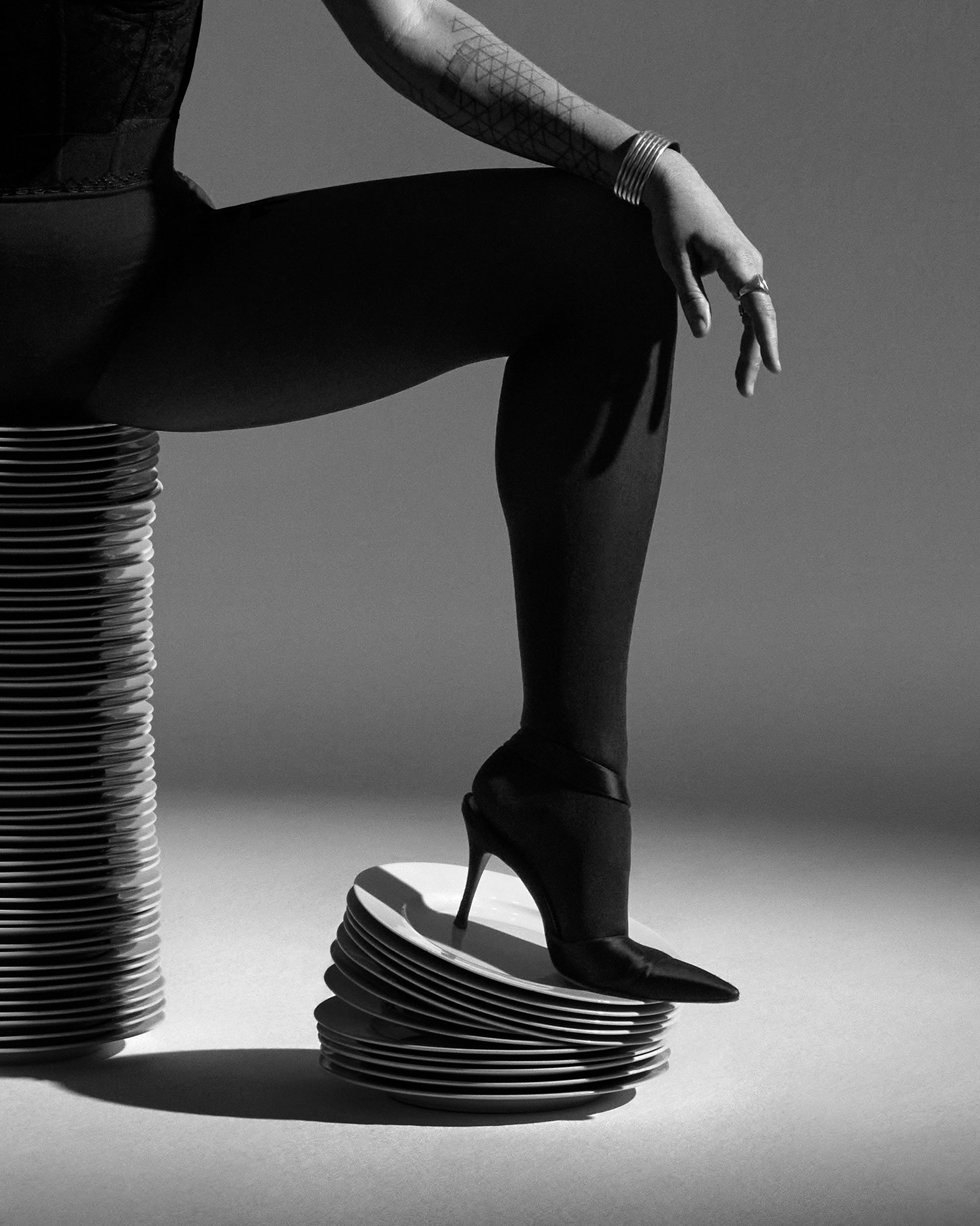Black and white image of a person’s leg in sheer tights and pointed high heel resting on stacked plates, with tattooed arm visible