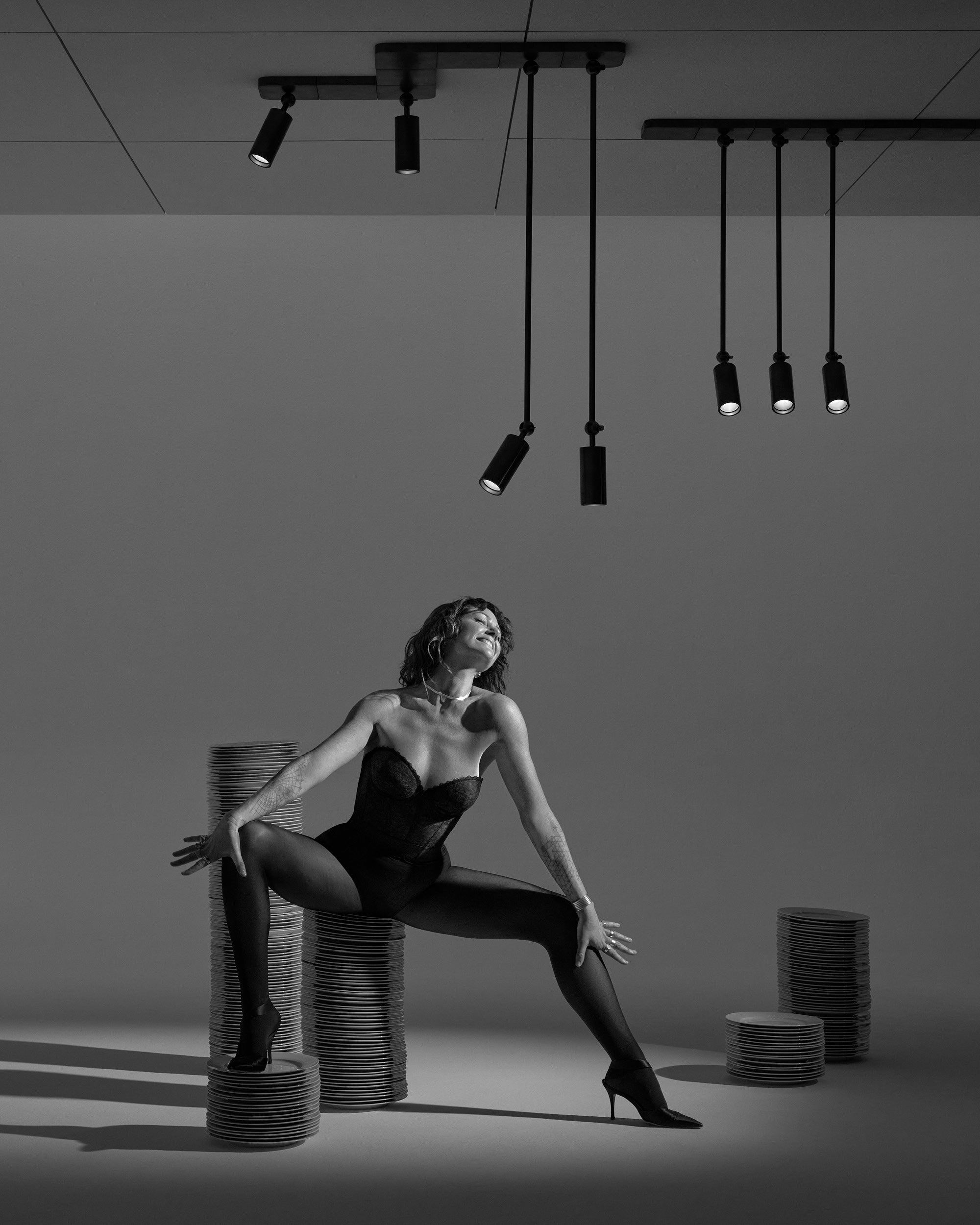 Black and white image of a woman in bodysuit and heels posing with stacks of round plates under pendant spotlights