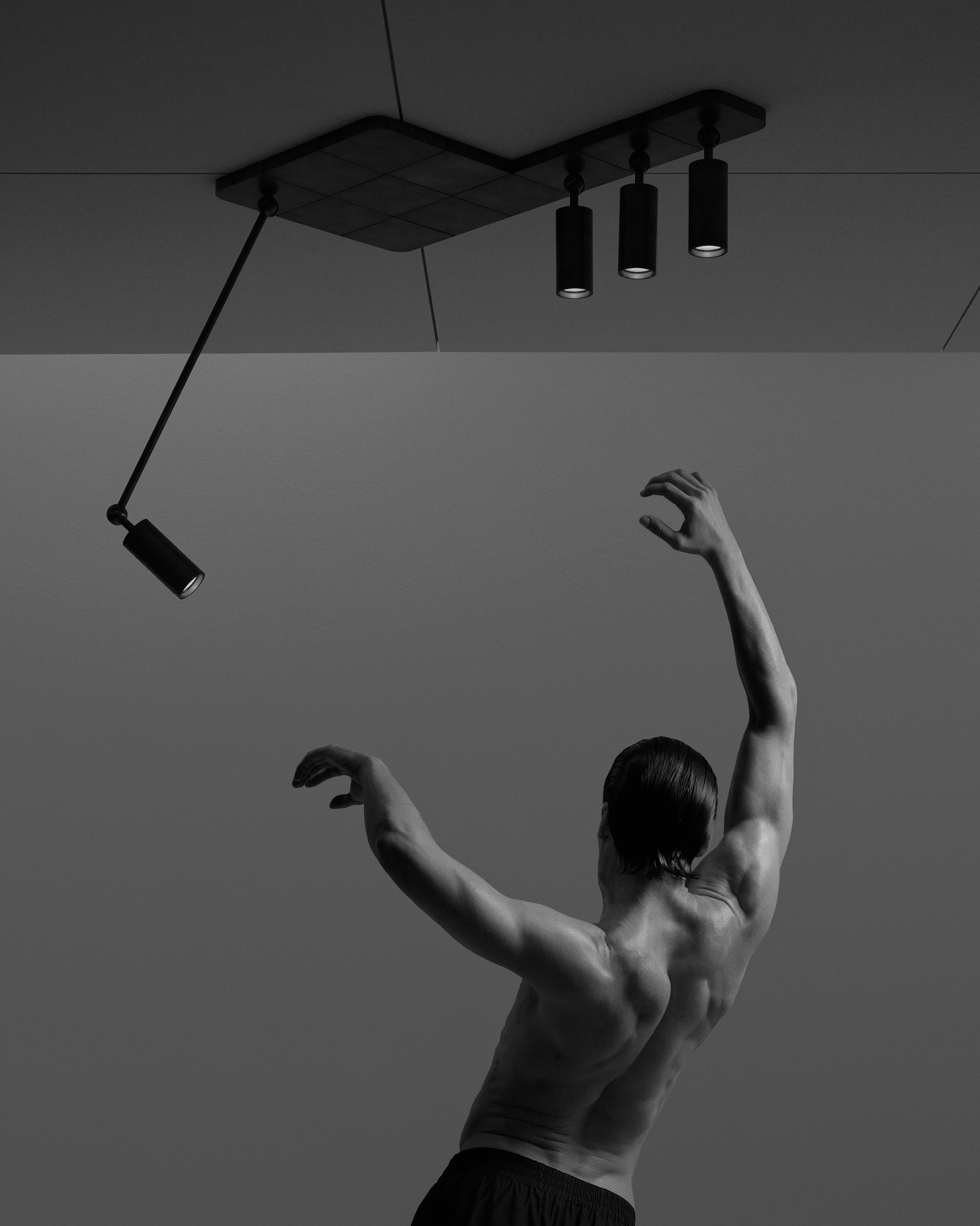 Black and white image of a shirtless male dancer with arms raised under modern ceiling spotlights
