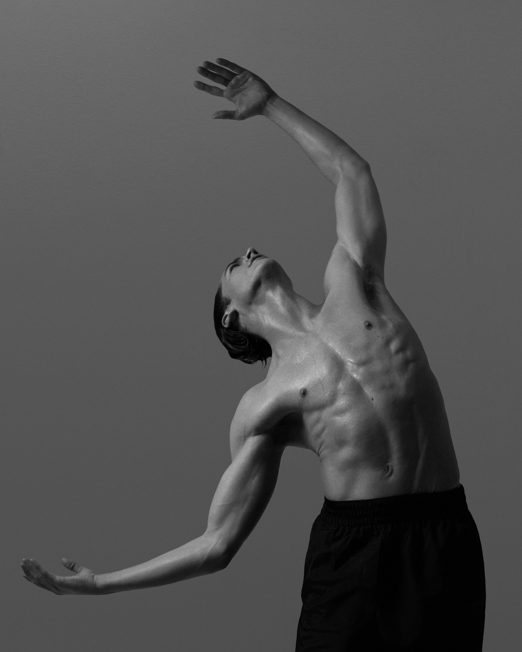 Black and white photo of a shirtless man in black pants posing with arms extended and head tilted back against plain background