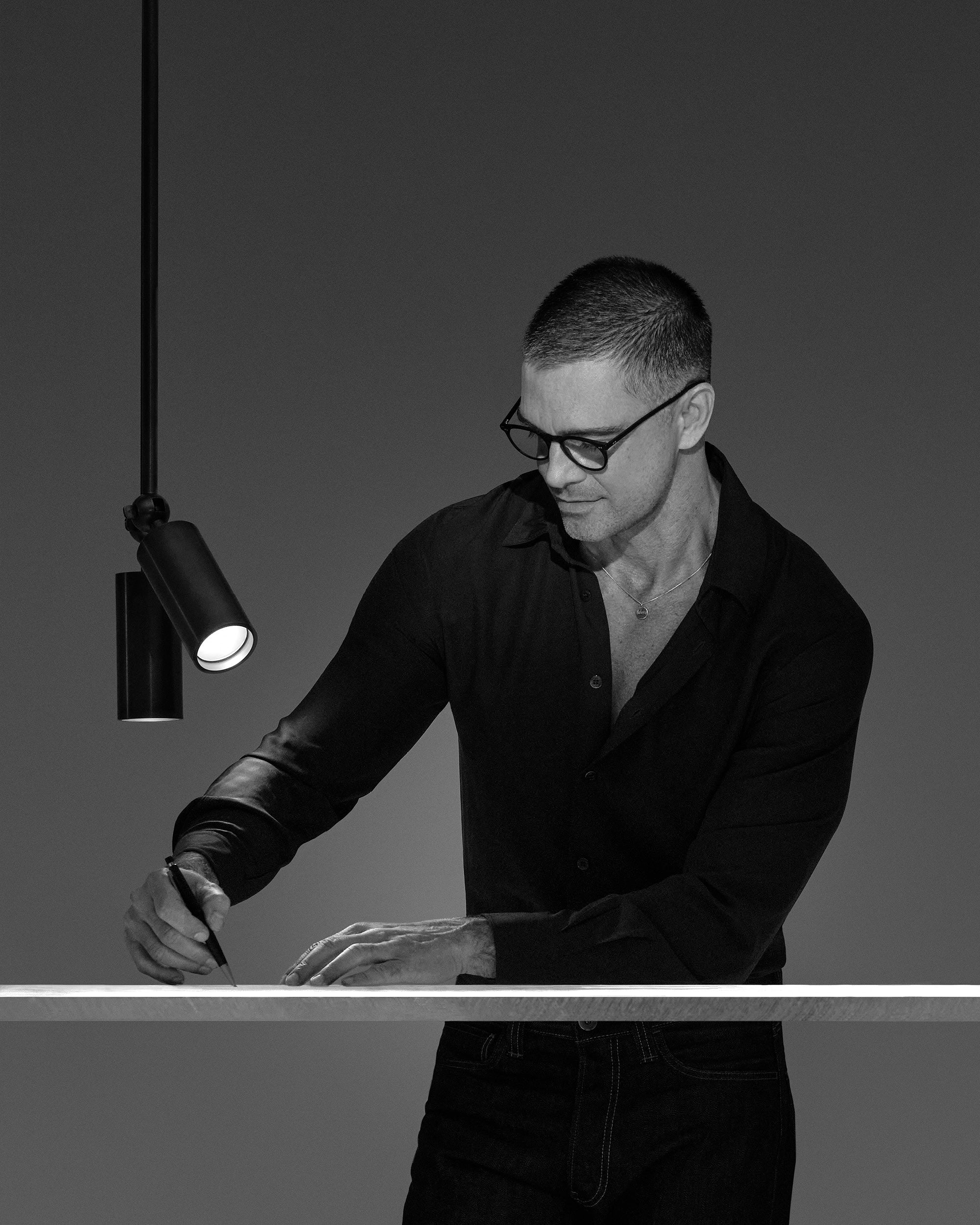 Black and white image of a man with glasses writing on a white surface under a modern hanging spotlight.