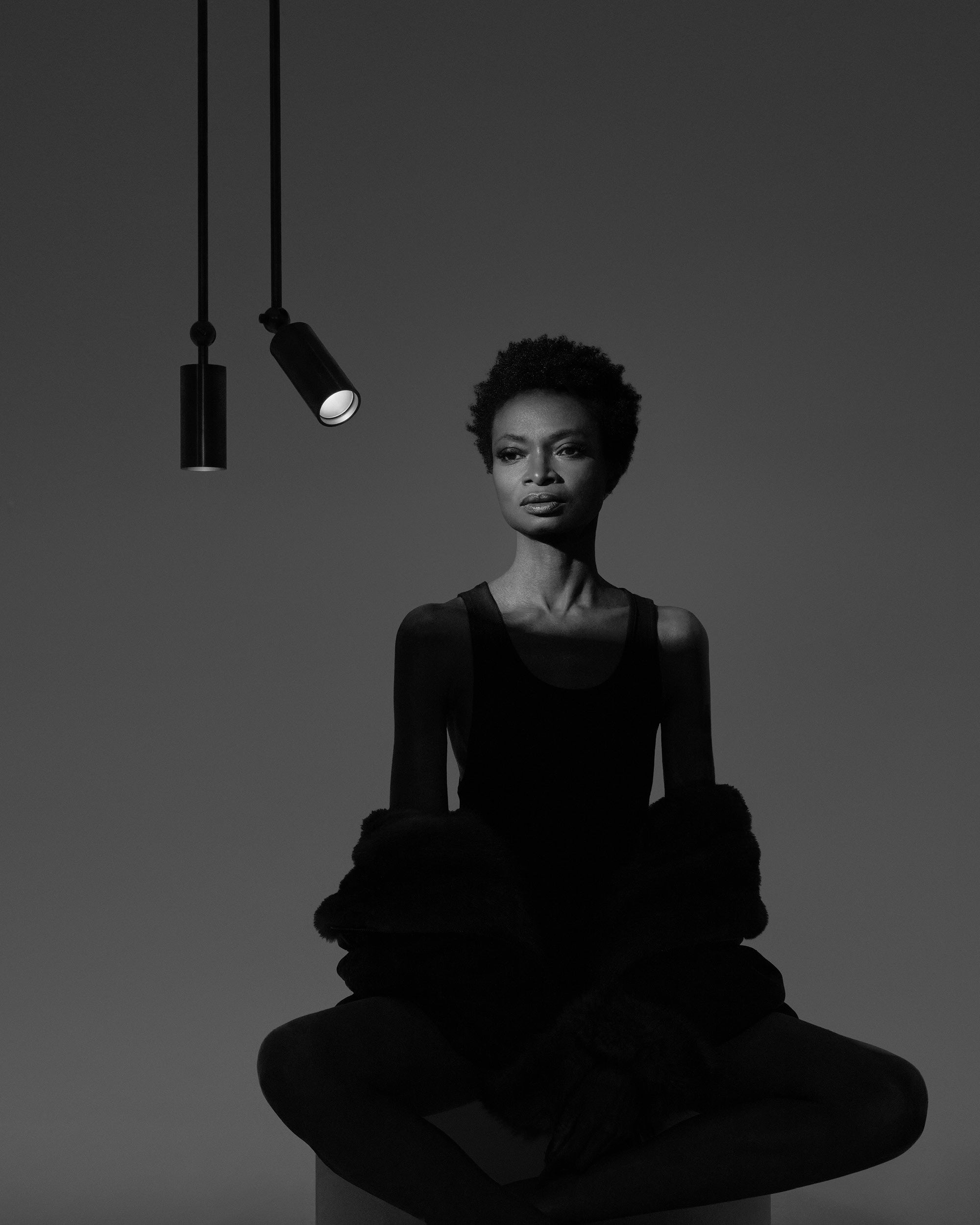 Black and white portrait of a woman seated cross-legged under two hanging spotlights against a plain background
