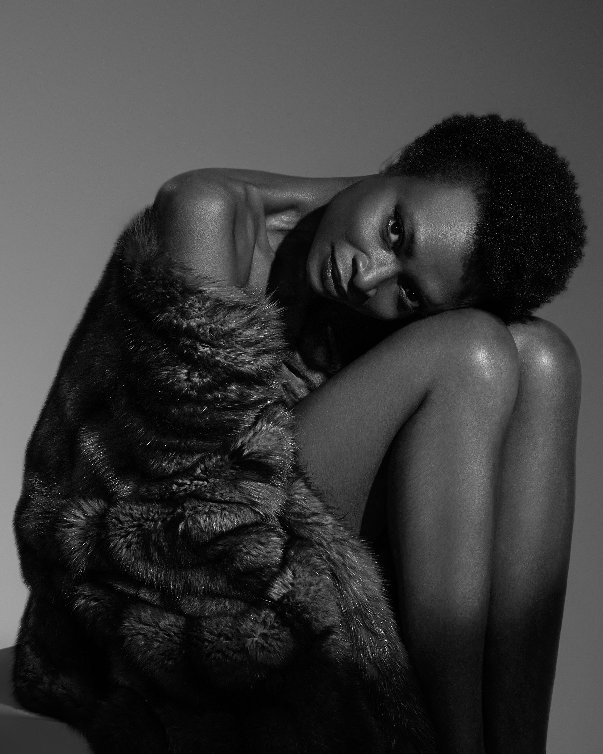Black and white portrait of a person with short hair wrapped in a textured fur coat, seated with knees drawn up.