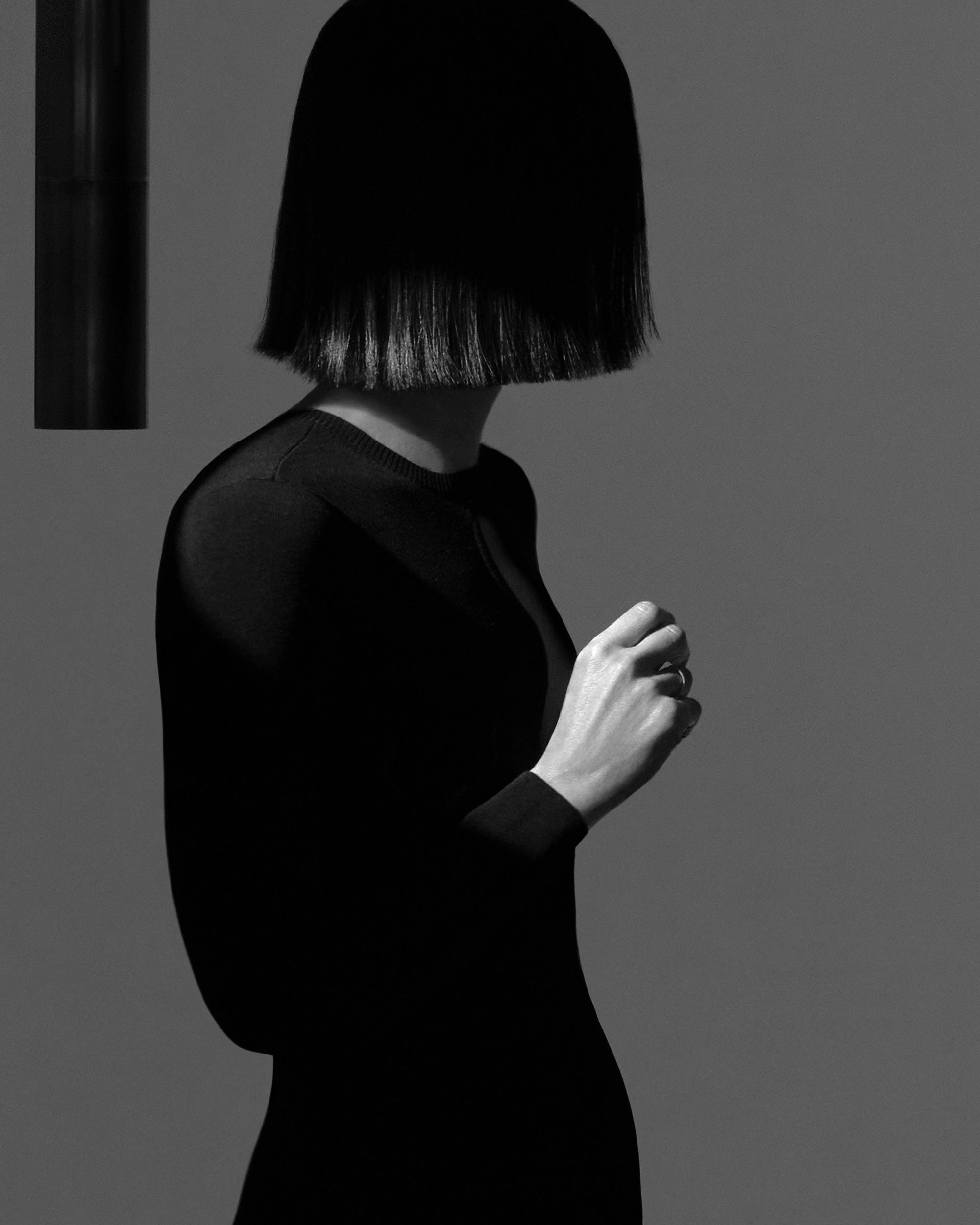 Black and white portrait of a person with straight hair covering the face, wearing a fitted long-sleeve top against a neutral background.