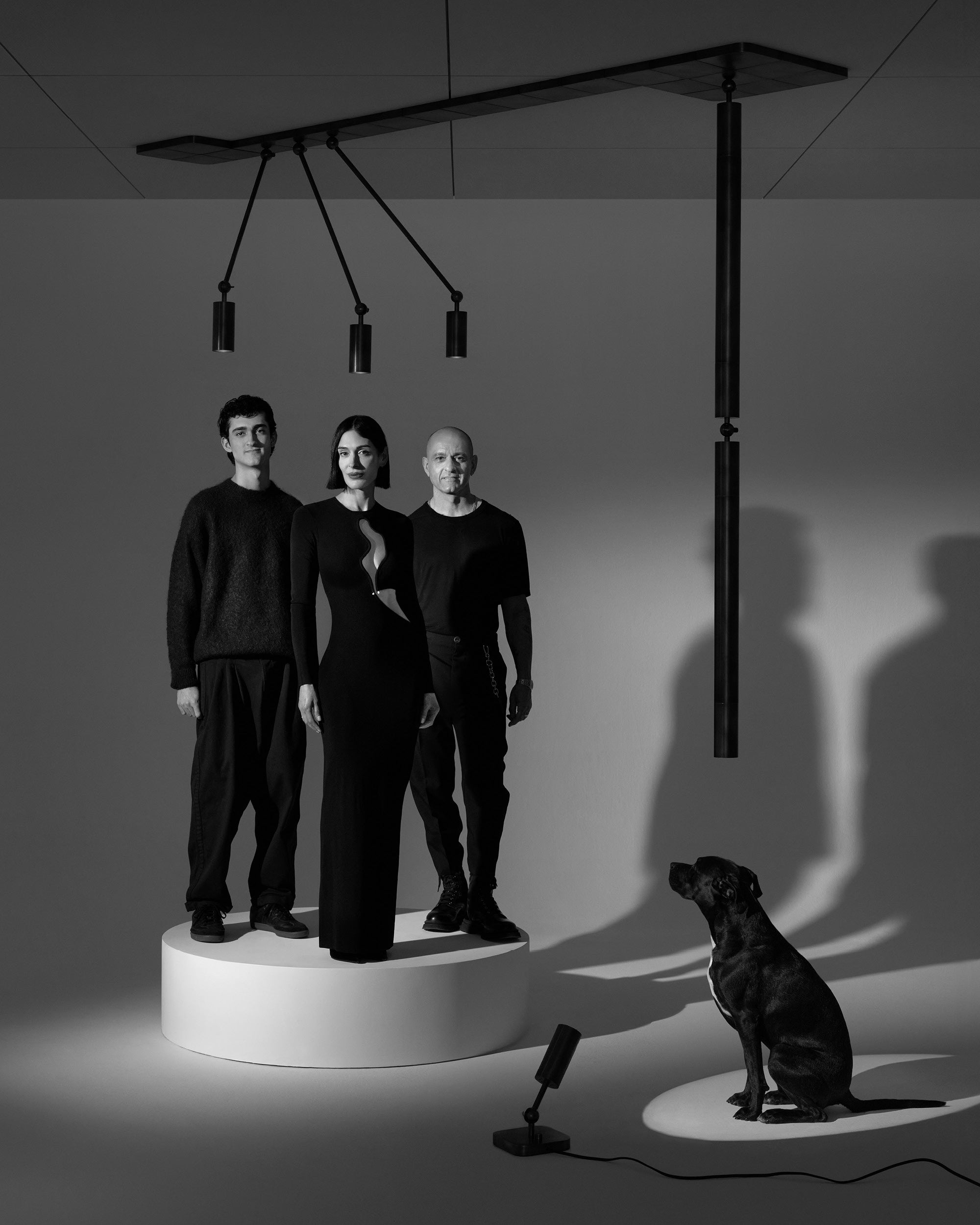 Black and white photo of three people standing on a white circular platform with a seated dog on a lit spot nearby and hanging lights above.
