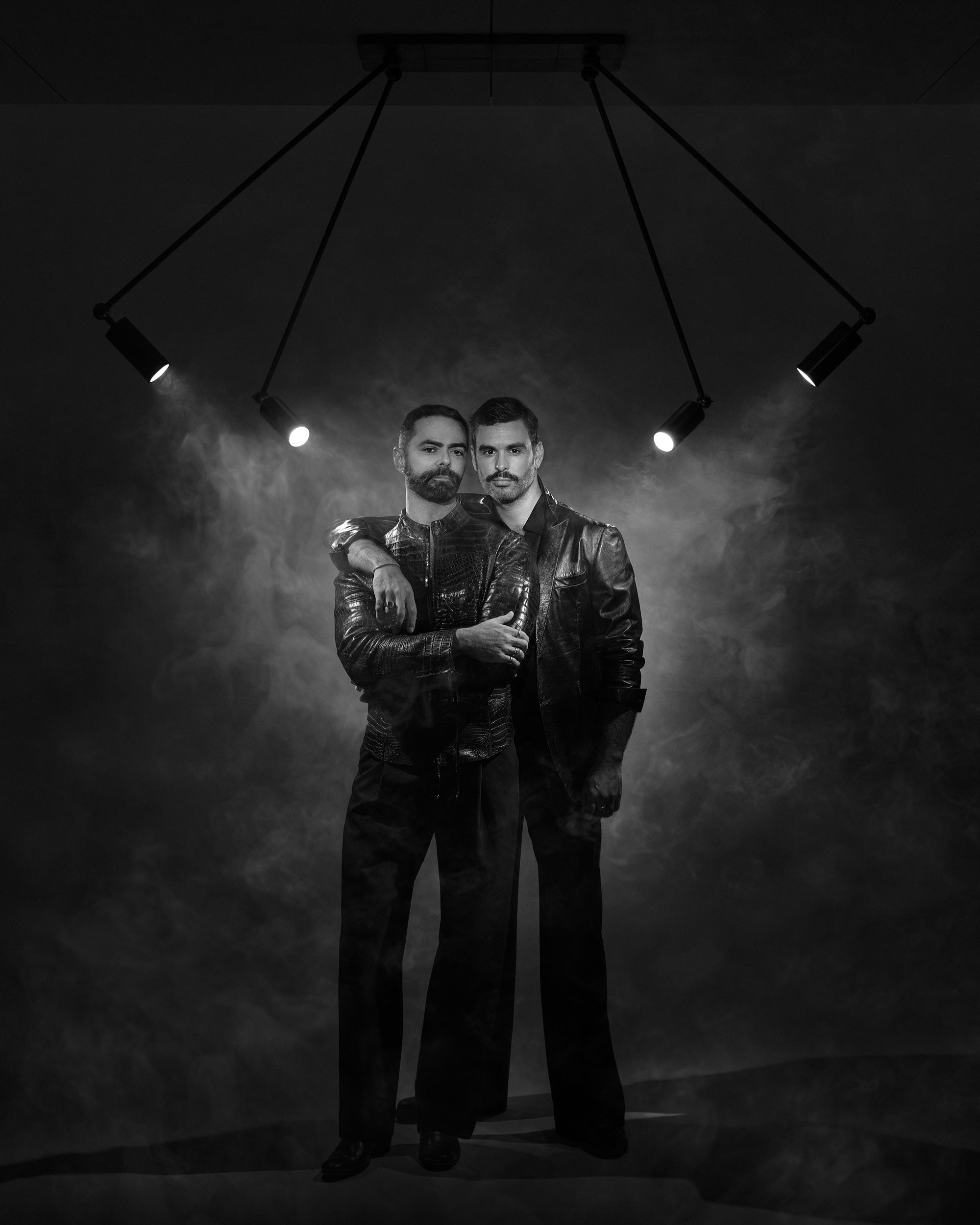 Black and white photo of two men in leather jackets standing closely under four angled spotlights with smoke in the background