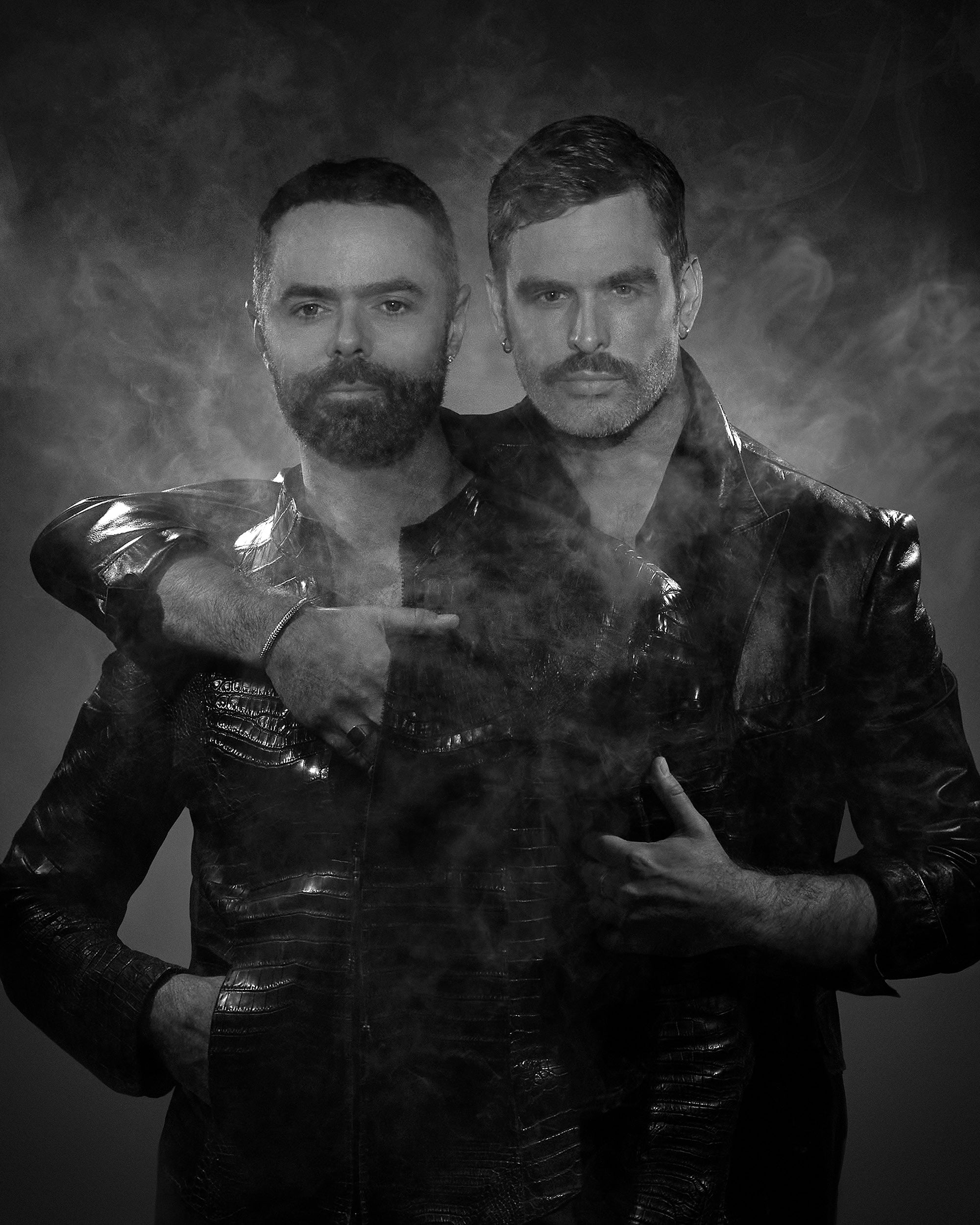 Black and white portrait of two men wearing textured leather jackets surrounded by swirling smoke against a dark background