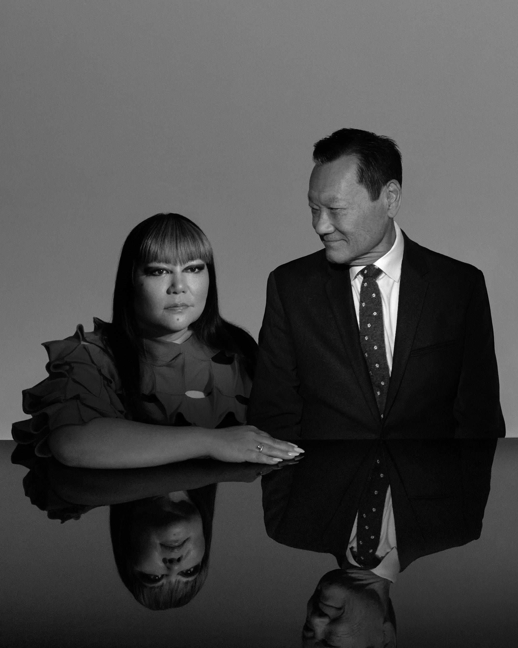 Black and white portrait of a woman and a man seated side by side with their reflections visible on a glossy surface.