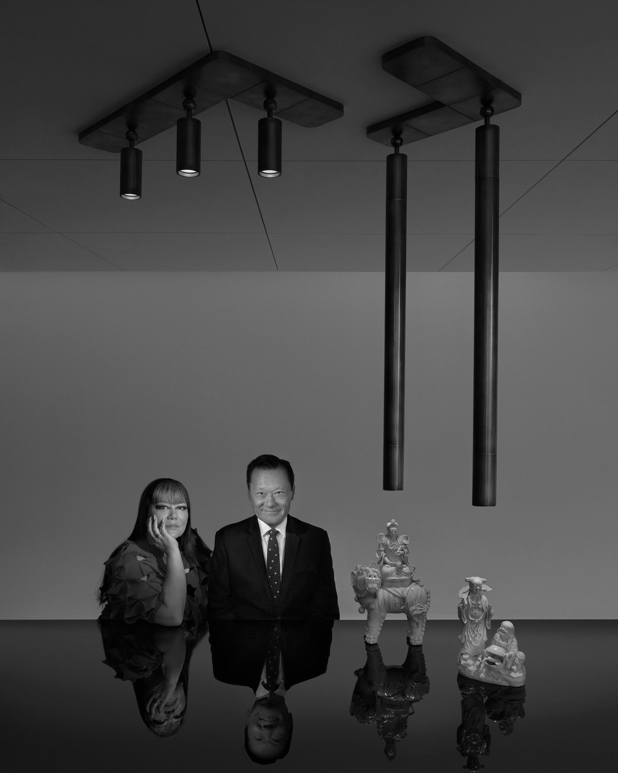 Black and white portrait of a man and woman seated at a reflective table with three sculptural figurines and ceiling lights above.