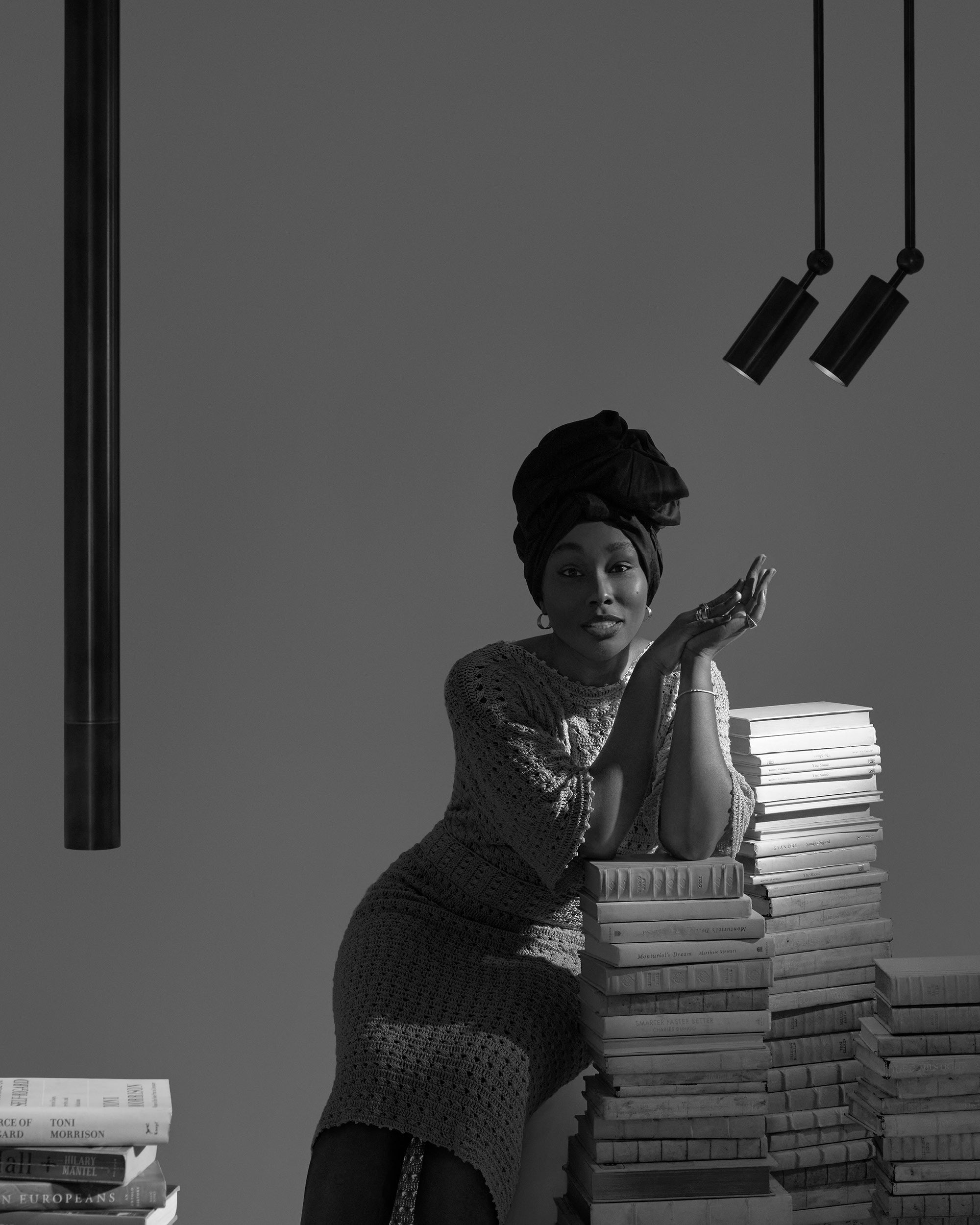 Black and white image of a woman wearing a headwrap leaning on stacks of books under two hanging cylindrical lights.