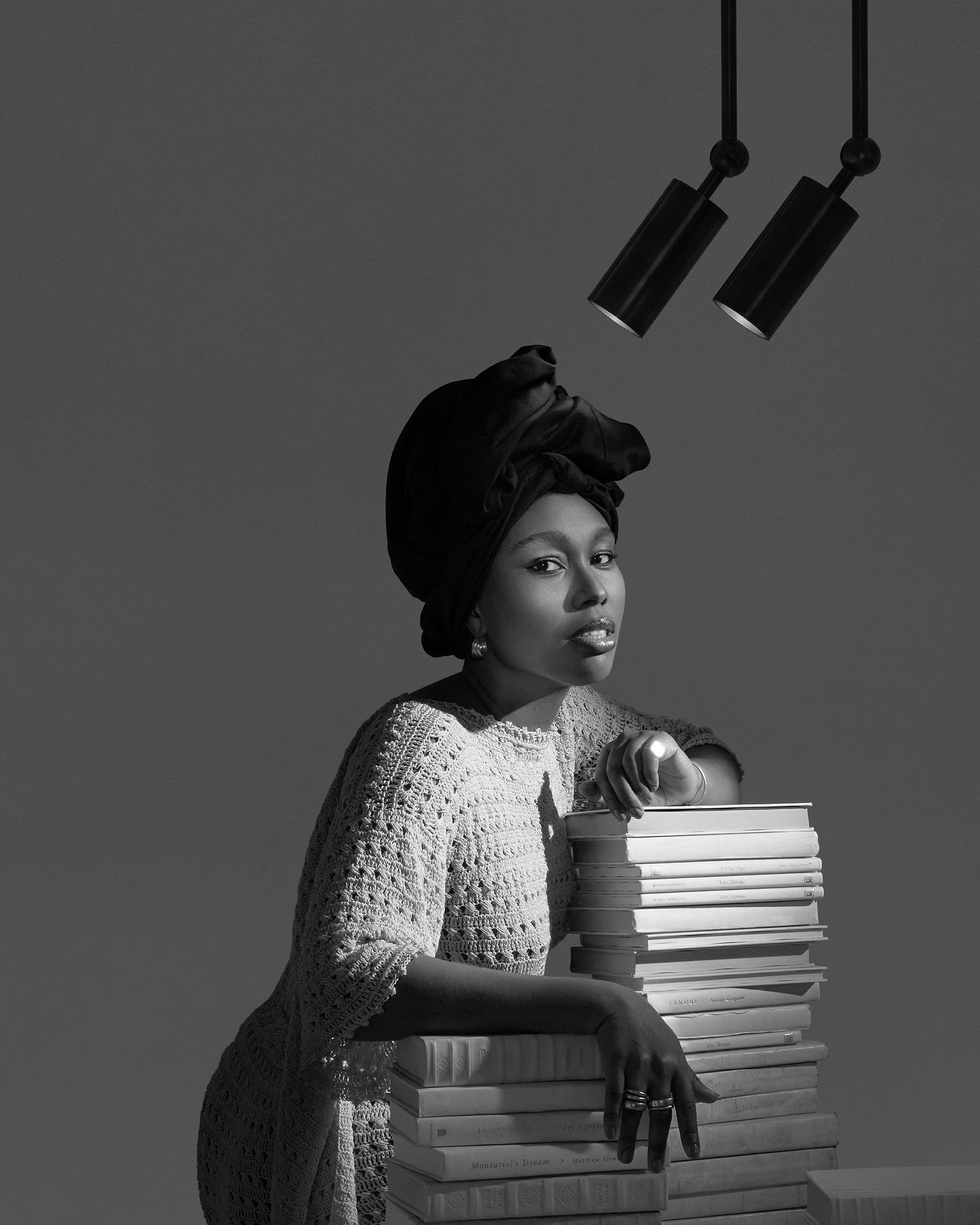 Black and white portrait of a woman wearing a headwrap leaning on a tall stack of books with two pendant lights above.
