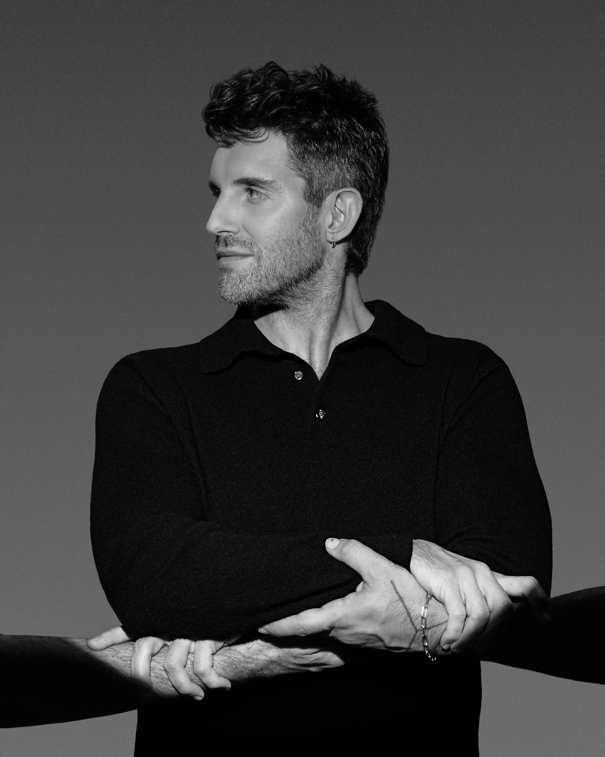 Black and white portrait of a man wearing a dark shirt, looking to the side with three hands holding his arms.
