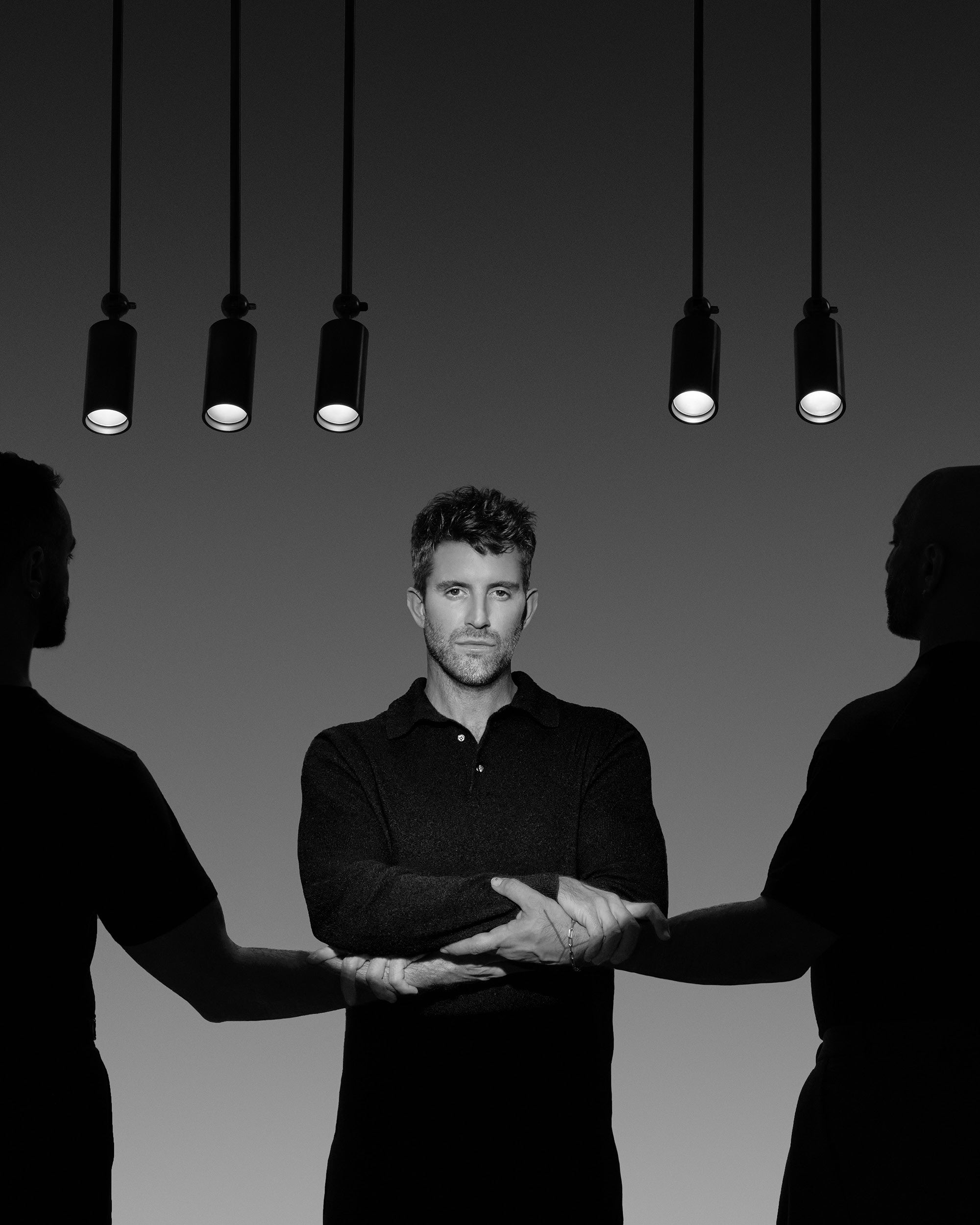 Black and white photo of a man illuminated by overhead spotlights, holding hands with two silhouetted figures on each side