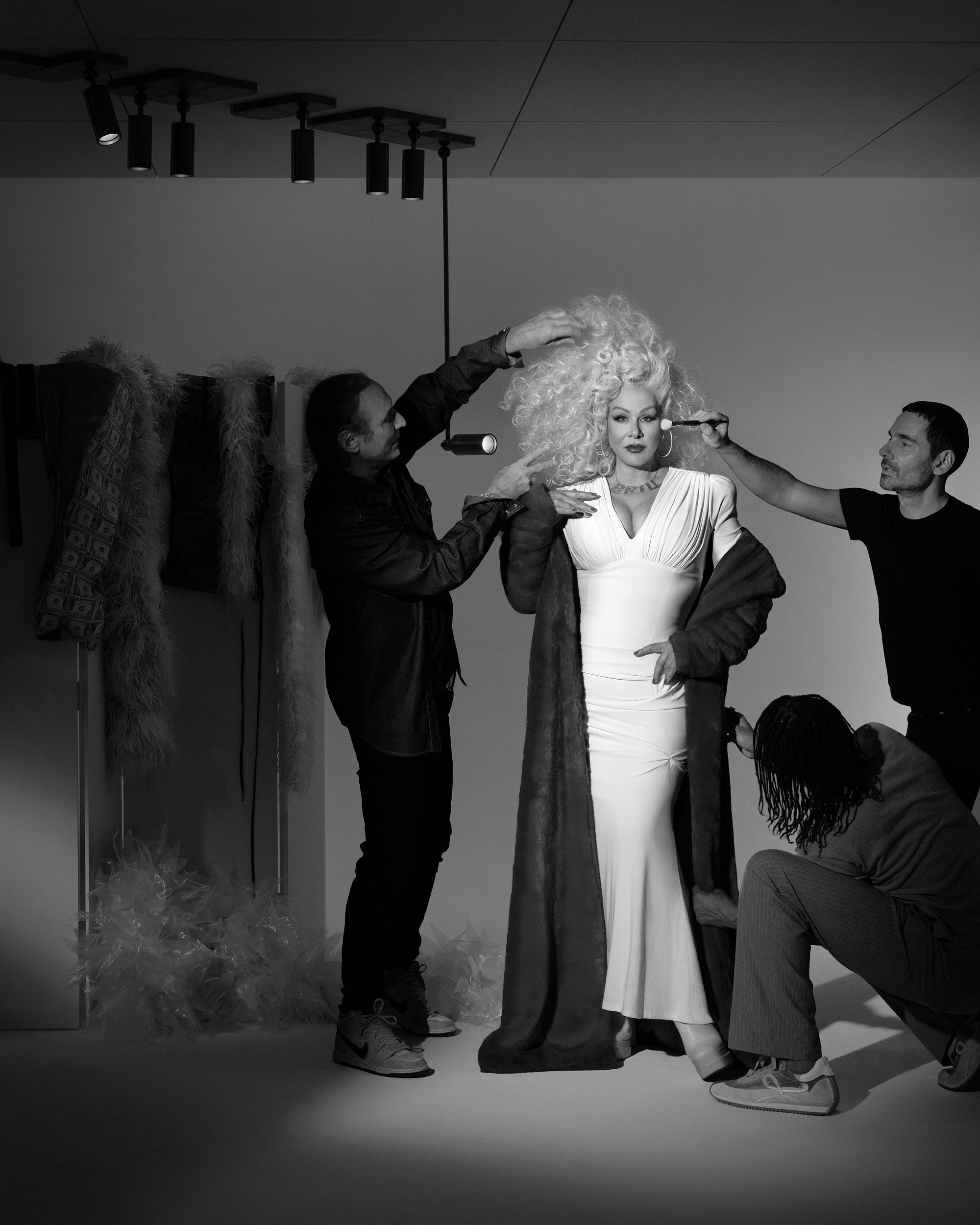 Black and white photo of a woman in a white dress and fur coat styled by three people adjusting her hair, makeup, and outfit