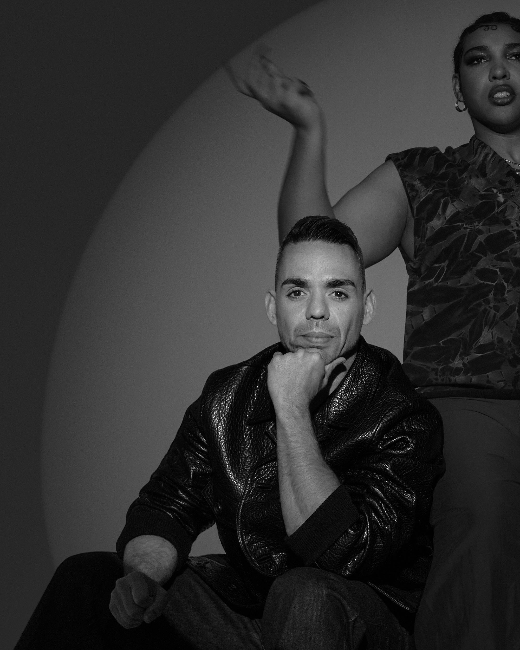 Black and white portrait of a man sitting with hand on chin and a partially visible person standing behind against a plain background