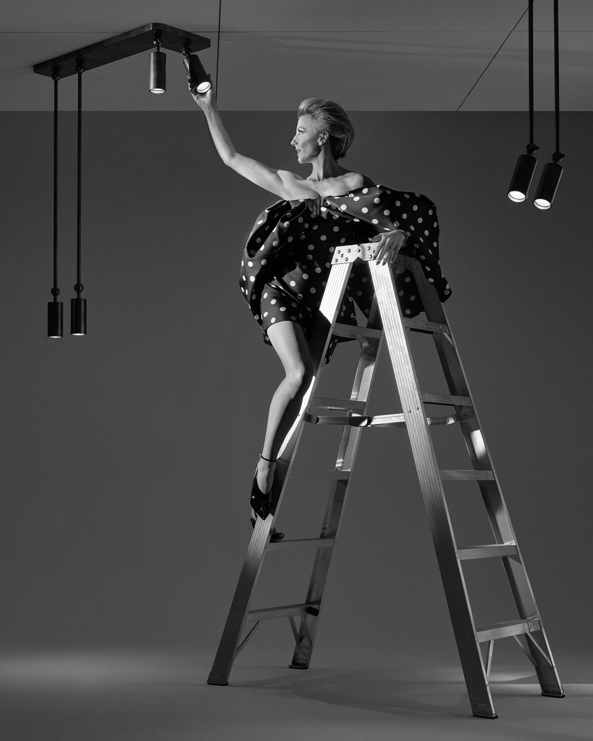 Black and white image of a woman in a polka dot dress adjusting a spotlight while standing on a metal ladder against a plain background